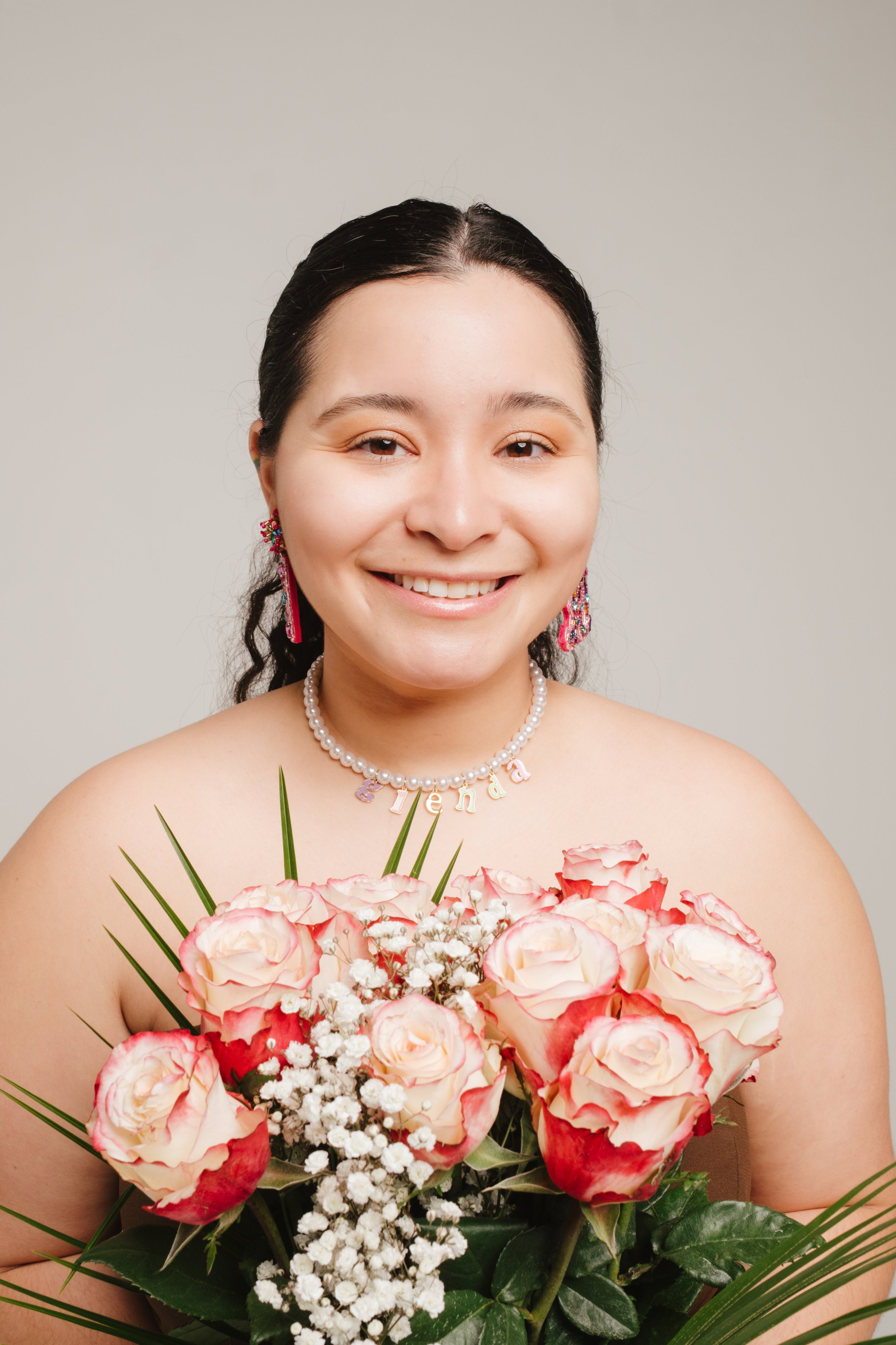 A woman with dark hair and light skin, smiling, holding a bouquet of pink and cream roses with white baby's breath and green leaves, wearing jewelry including a necklace with colorful letters spelling 'Freda' and earrings with pink and purple beads, 
