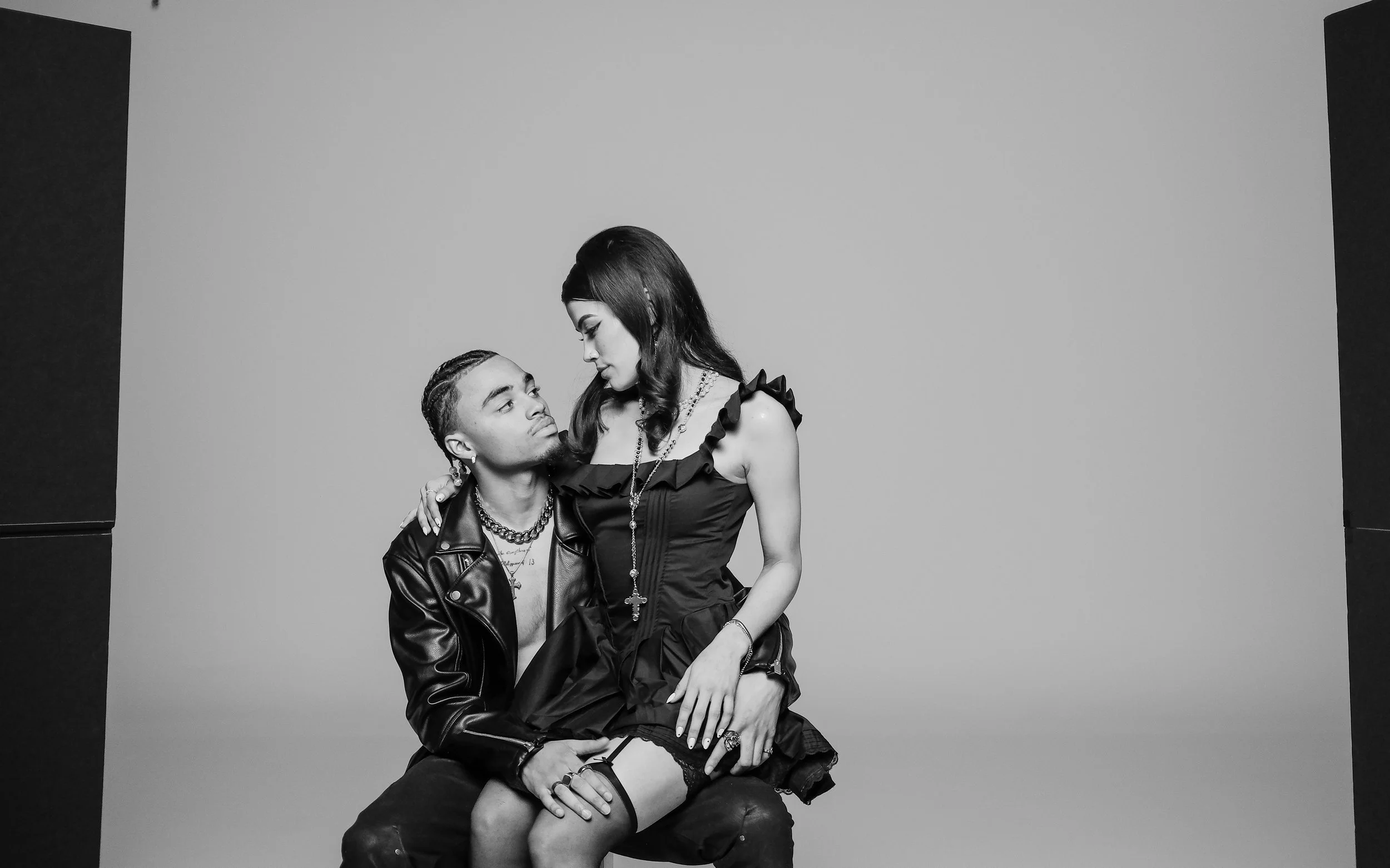 Black and white photo of a man and a woman in a studio. The man with braided hair, wearing a leather jacket and layered necklaces, is sitting. The woman, with long dark hair, is dressed in a ruffled dress and tall stockings, sitting on the man's lap 