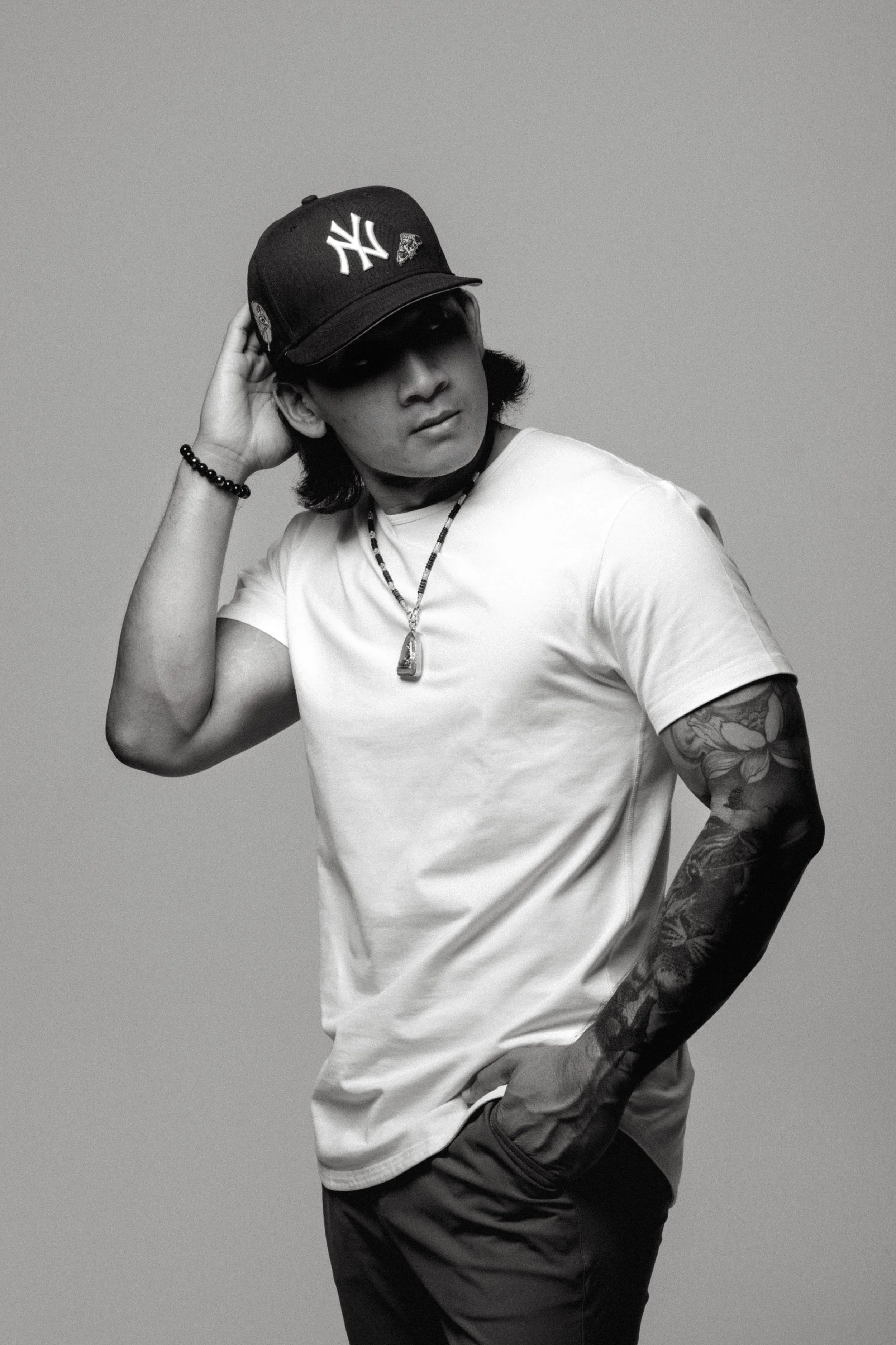 A young man with shoulder-length hair wearing a New York Yankees cap, white t-shirt, necklace, wristband, and tattooed arm, posing against a plain background.