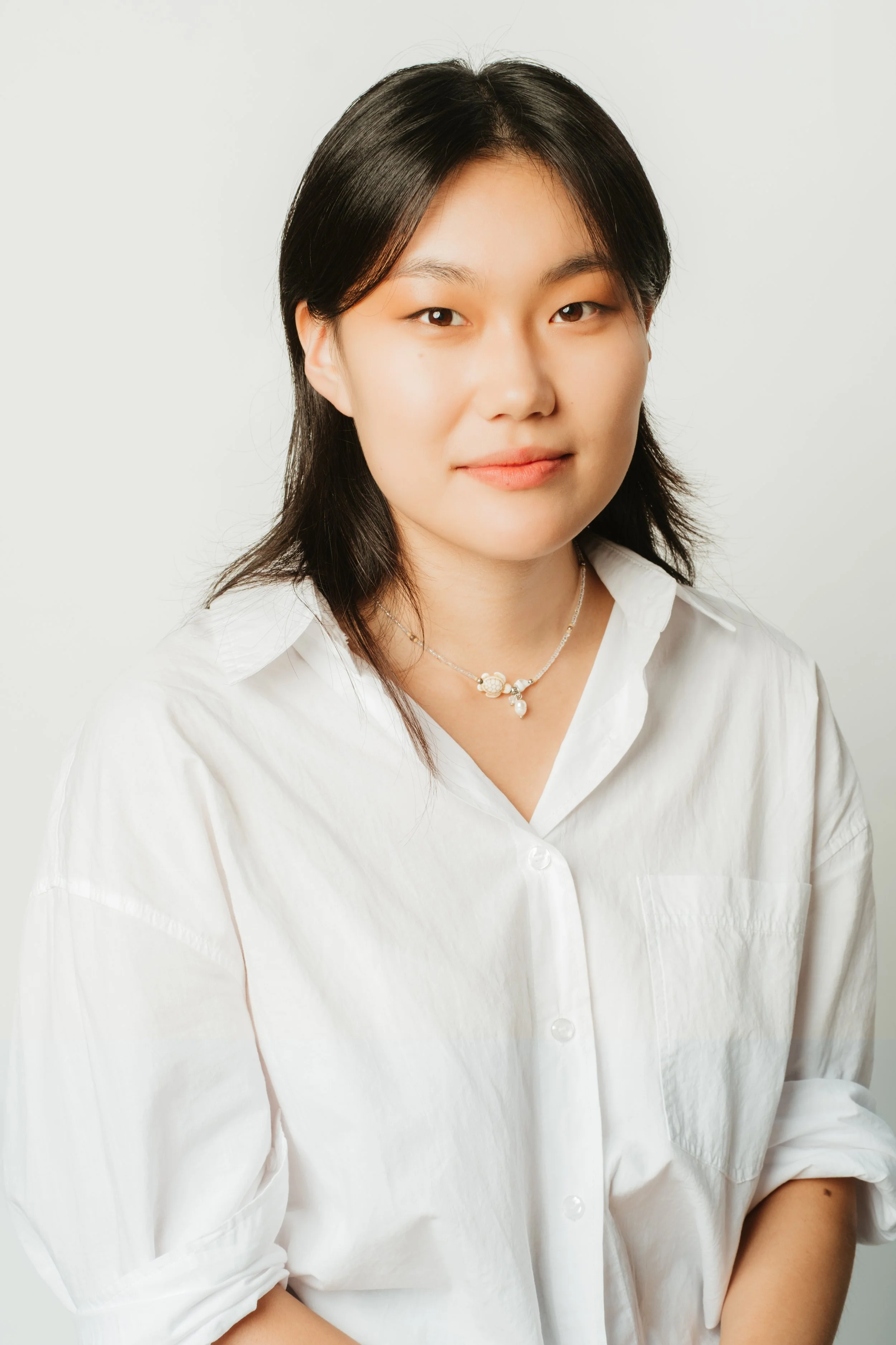Portrait of a young woman with black hair in a white shirt and necklace against a plain background.