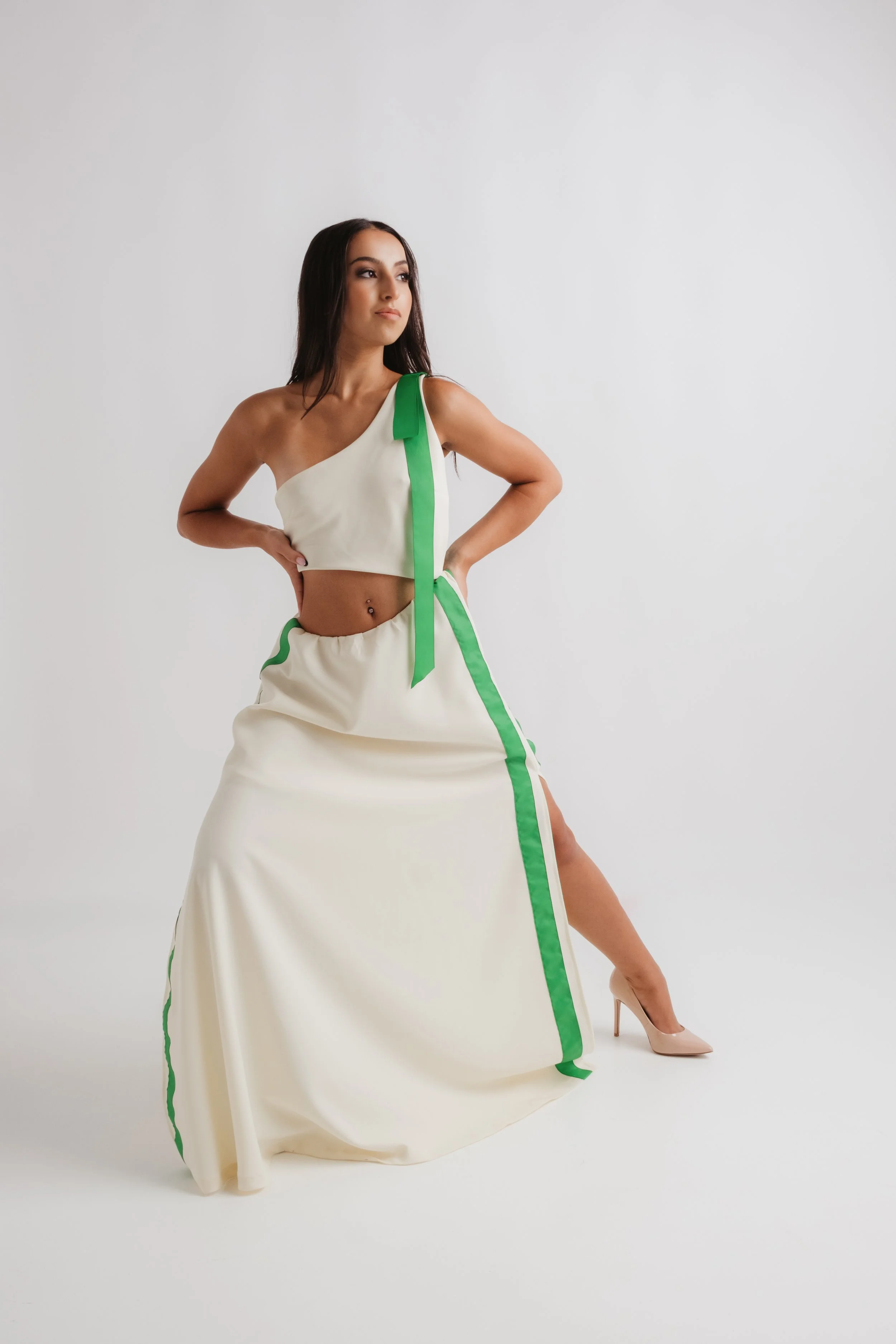 A woman in a cream-colored, two-piece outfit with green accents, standing with one hand on her hip and the other resting on her waist, wearing nude high heels against a plain white background.