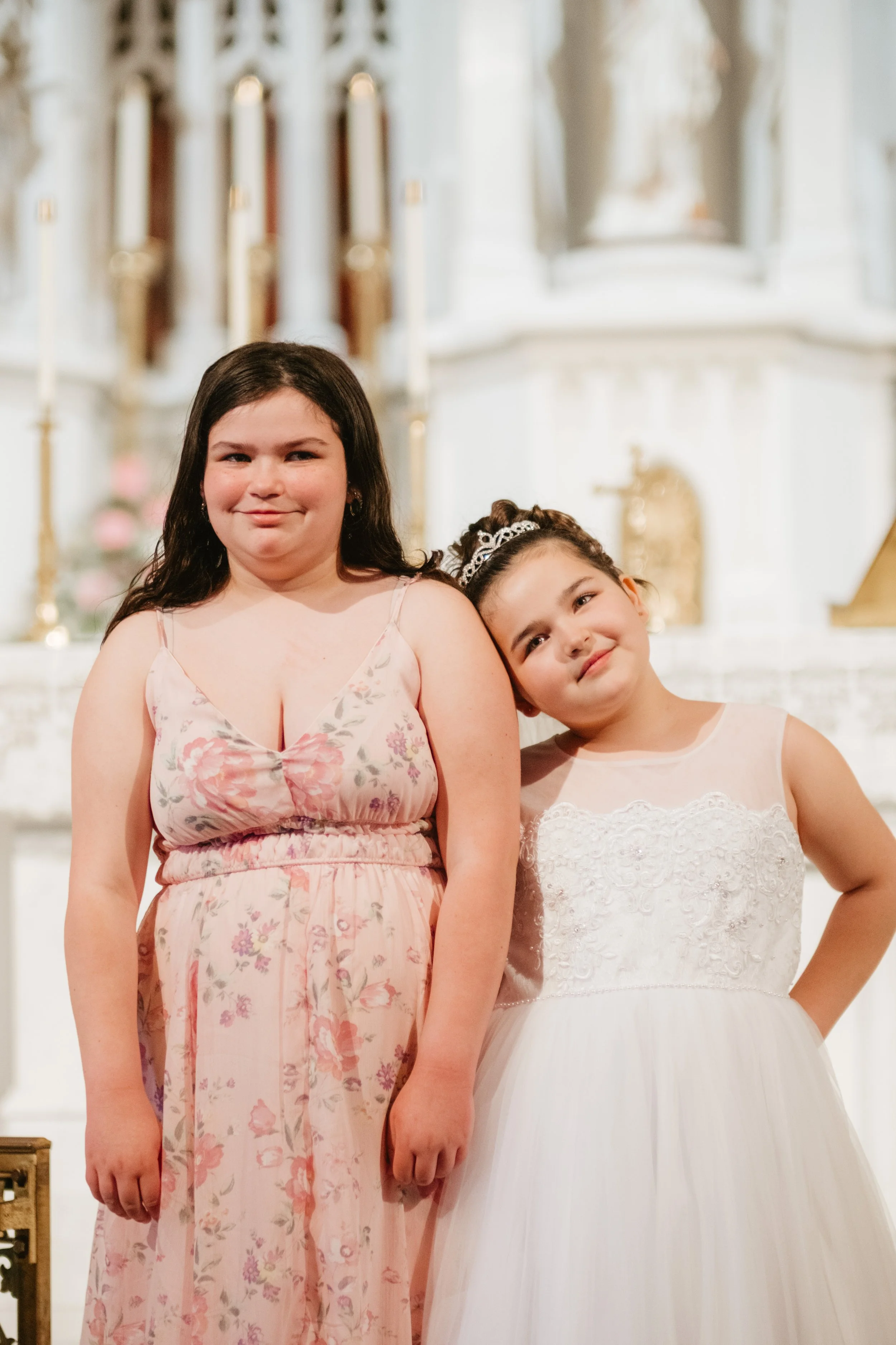 Two young girls in dresses standing inside a church or cathedral with candles and religious decor in the background. They appear to be at a special event, possibly a First Communion or similar celebration.