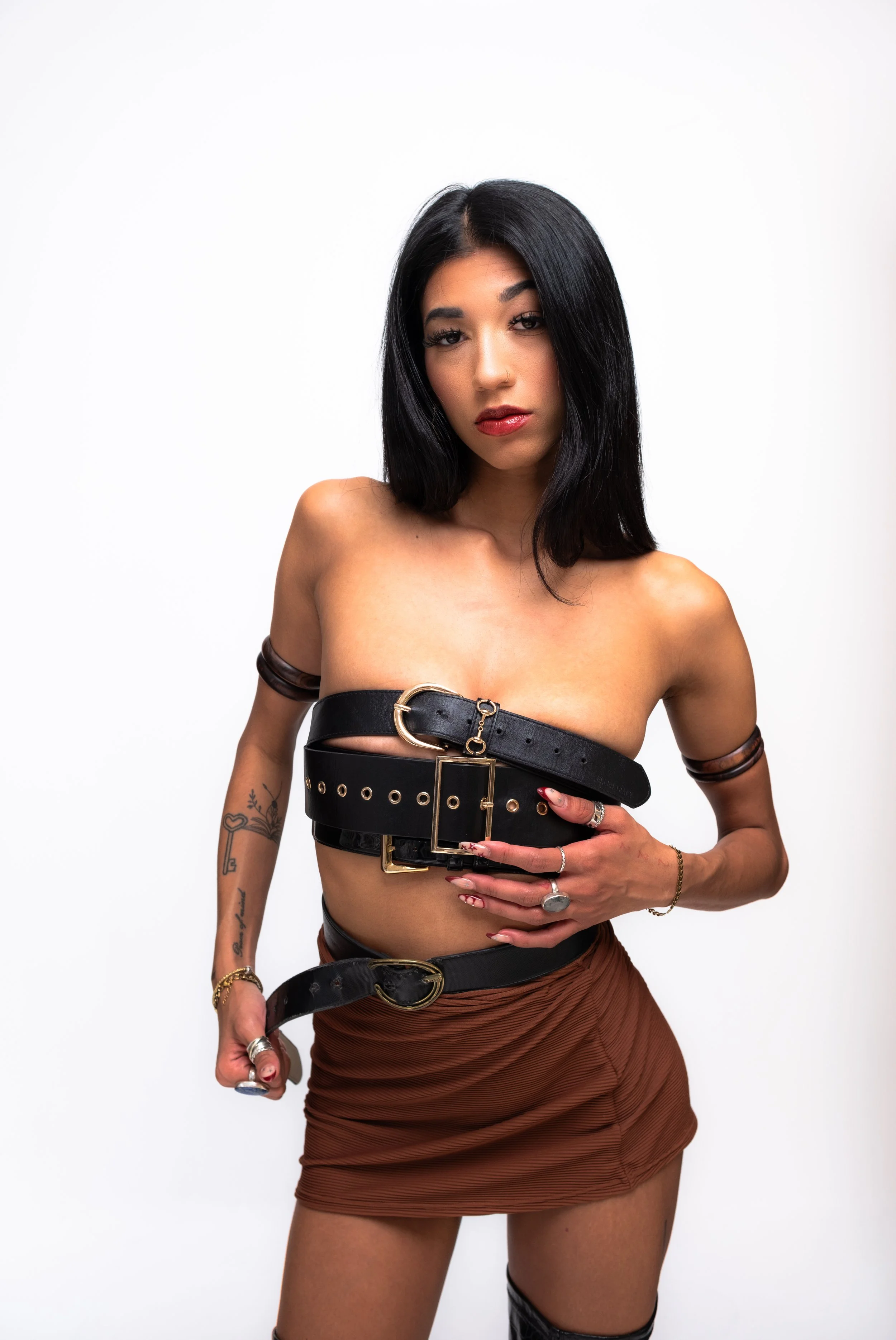 A woman with black hair and tattoos, wearing a black belt and brown skirt, standing against a plain white background.