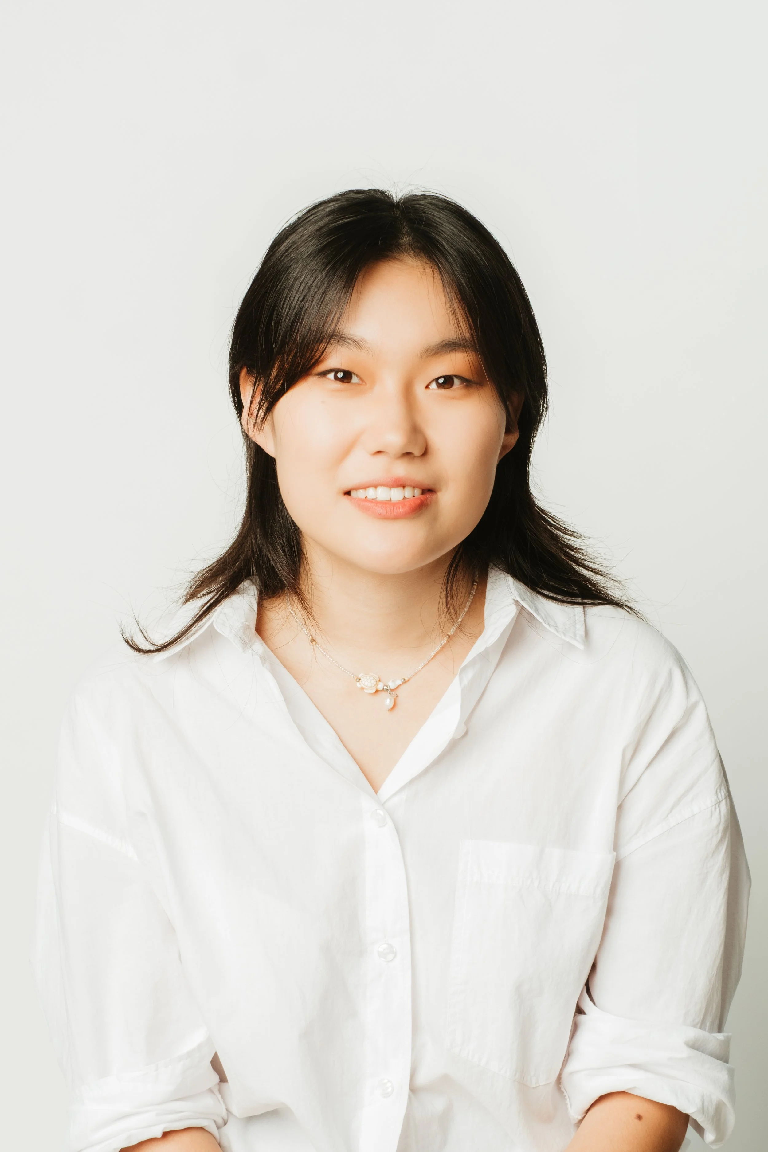 A young woman with black hair wearing a white button-up shirt and a necklace, smiling against a plain white background.