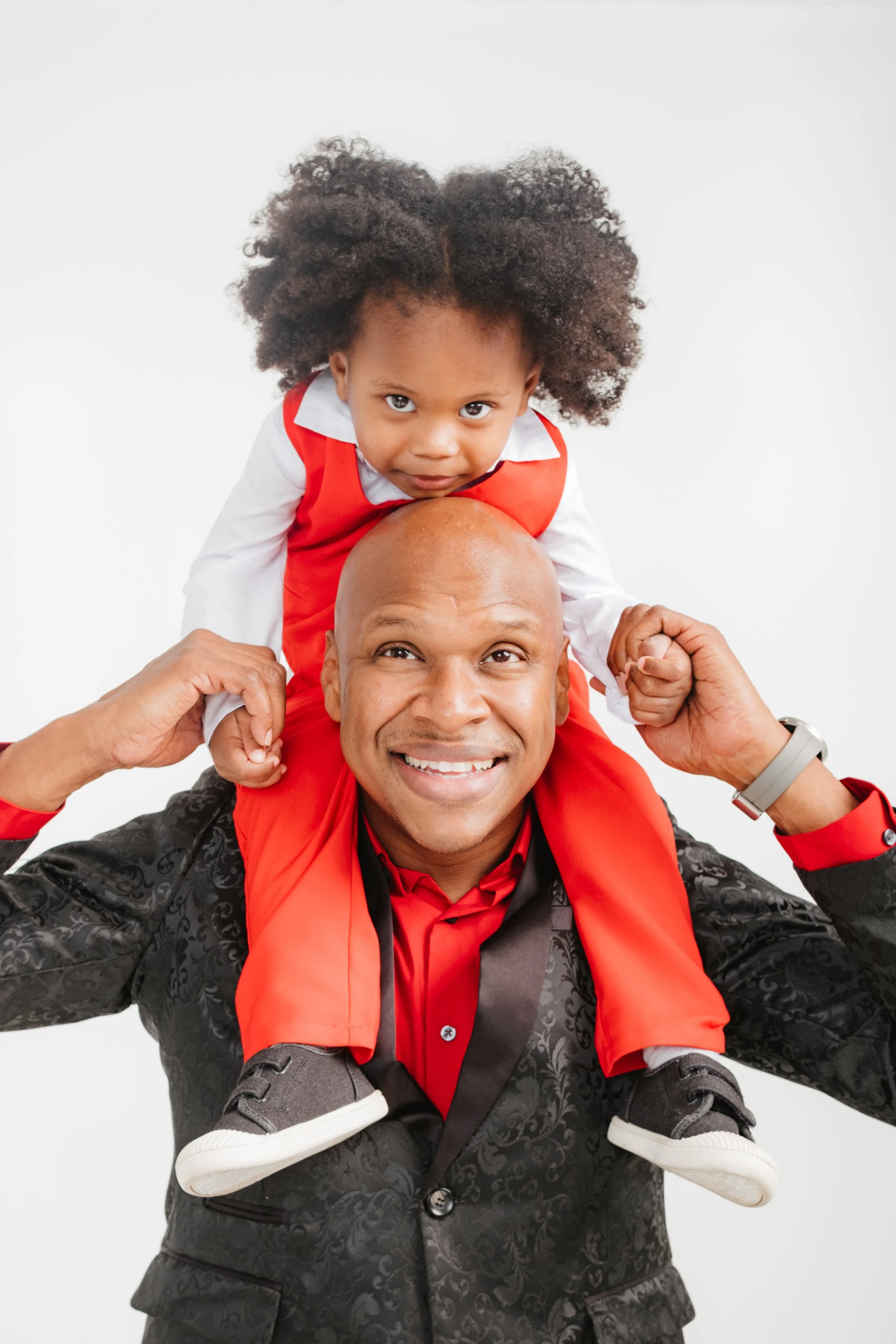 A man in a patterned black blazer, red shirt, and gray watch carries a young girl on his shoulders. The girl is wearing a red vest, white blouse, and black shoes. Both are smiling while the girl looks directly at the camera.
