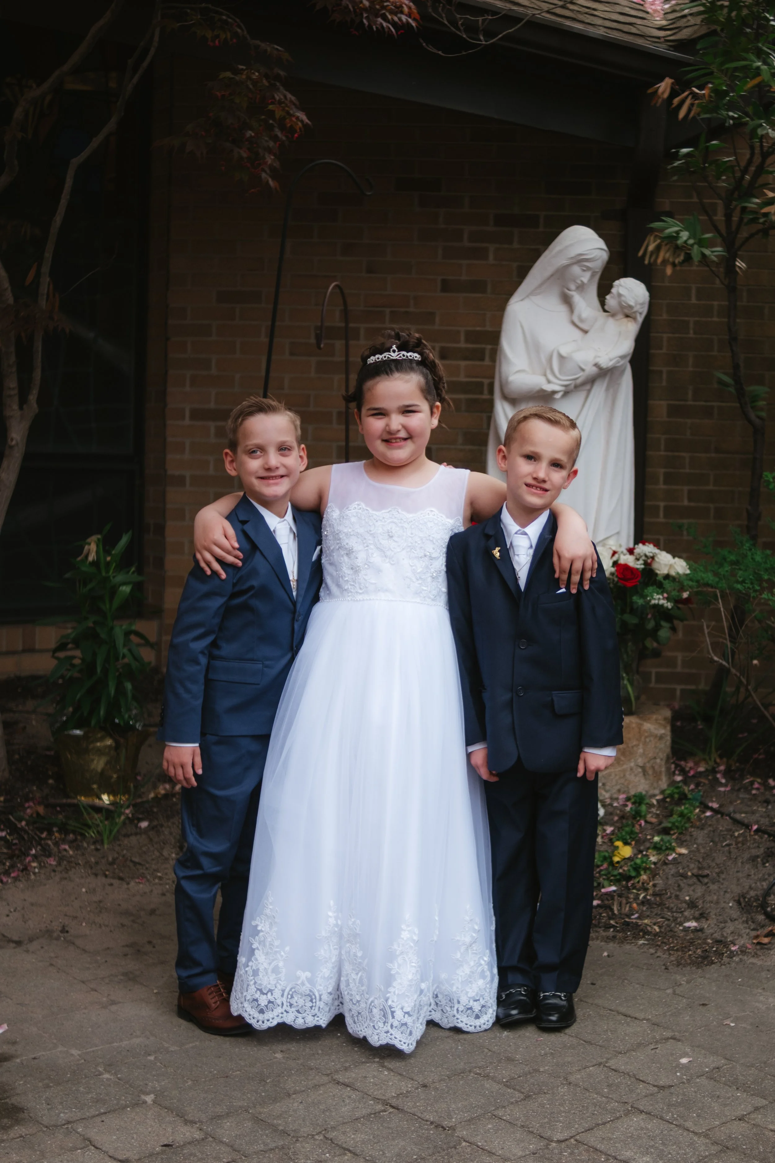 Three children standing outdoors in front of a religious statue. The girl in the center is wearing a white dress with lace details, and the two boys on either side are in dark suits.