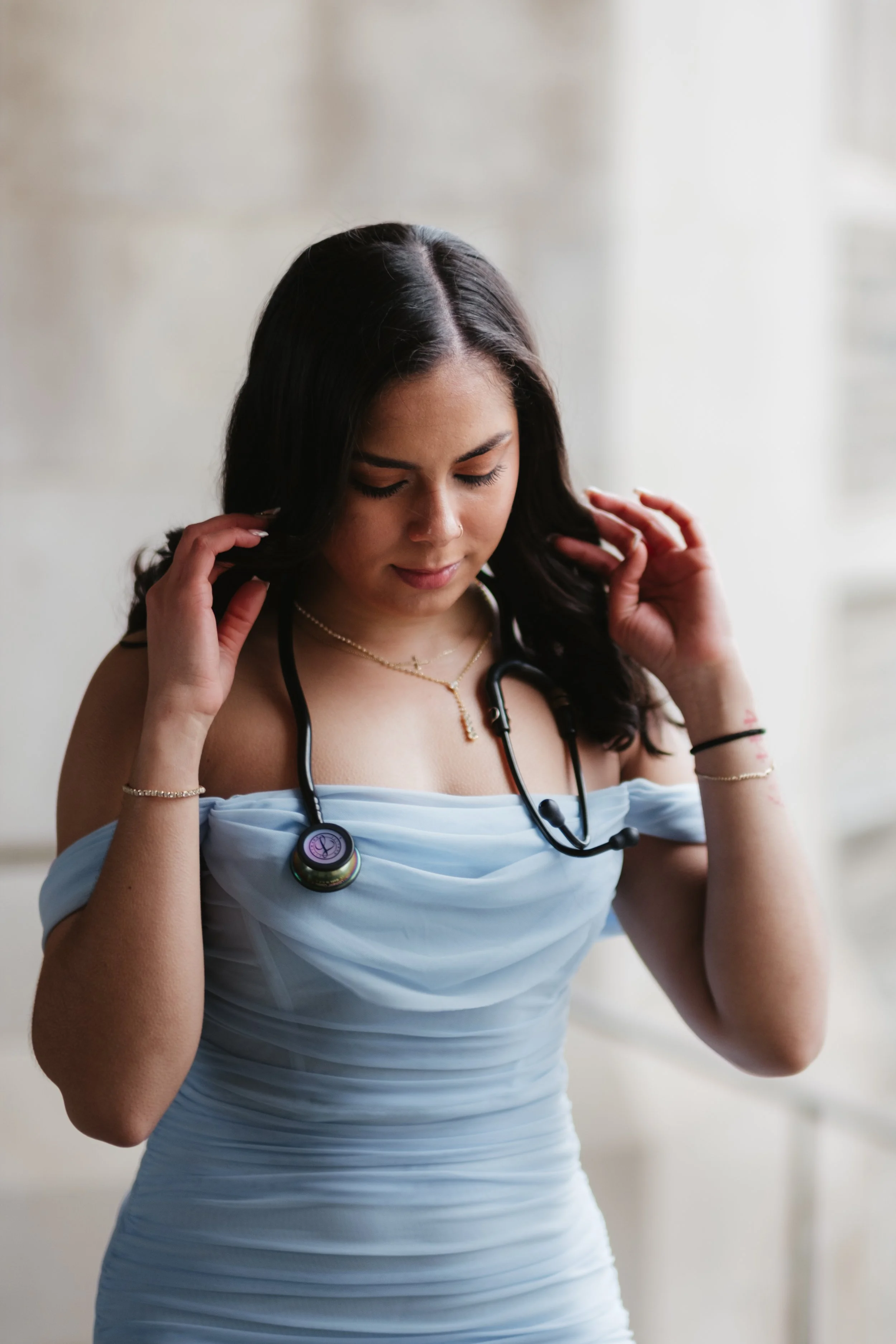 A young woman with dark hair, wearing a light blue off-shoulder dress, is adjusting her stethoscope around her neck with her eyes closed, standing in front of a light background.