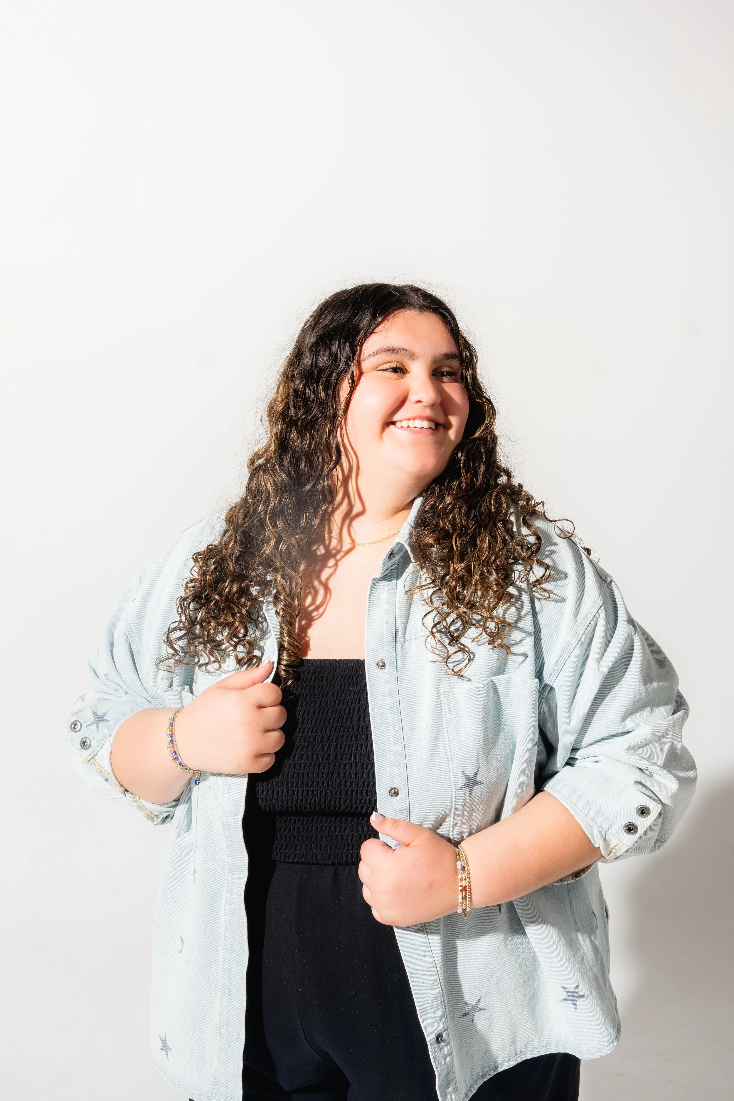 A smiling young woman with long, curly hair wearing a denim shirt with star patterns over a black top, standing against a white background.