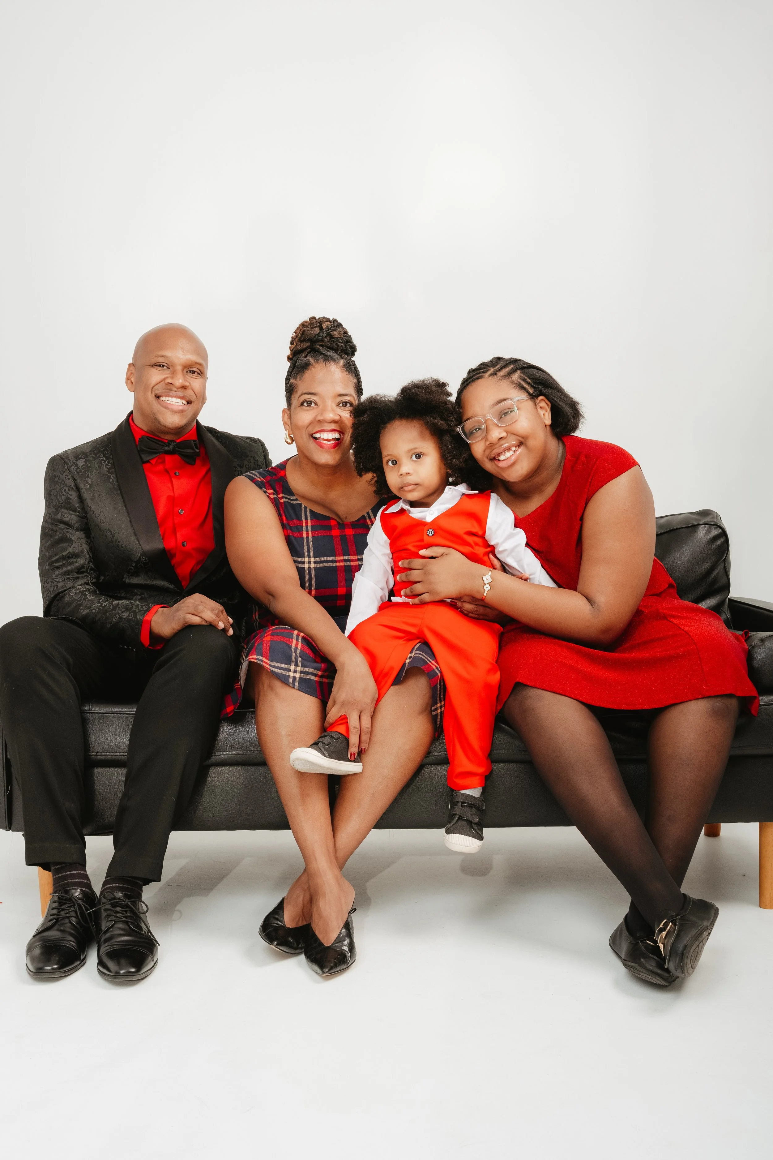 Family of four sitting on a black sofa, smiling, dressed in red and black outfits against a white background.