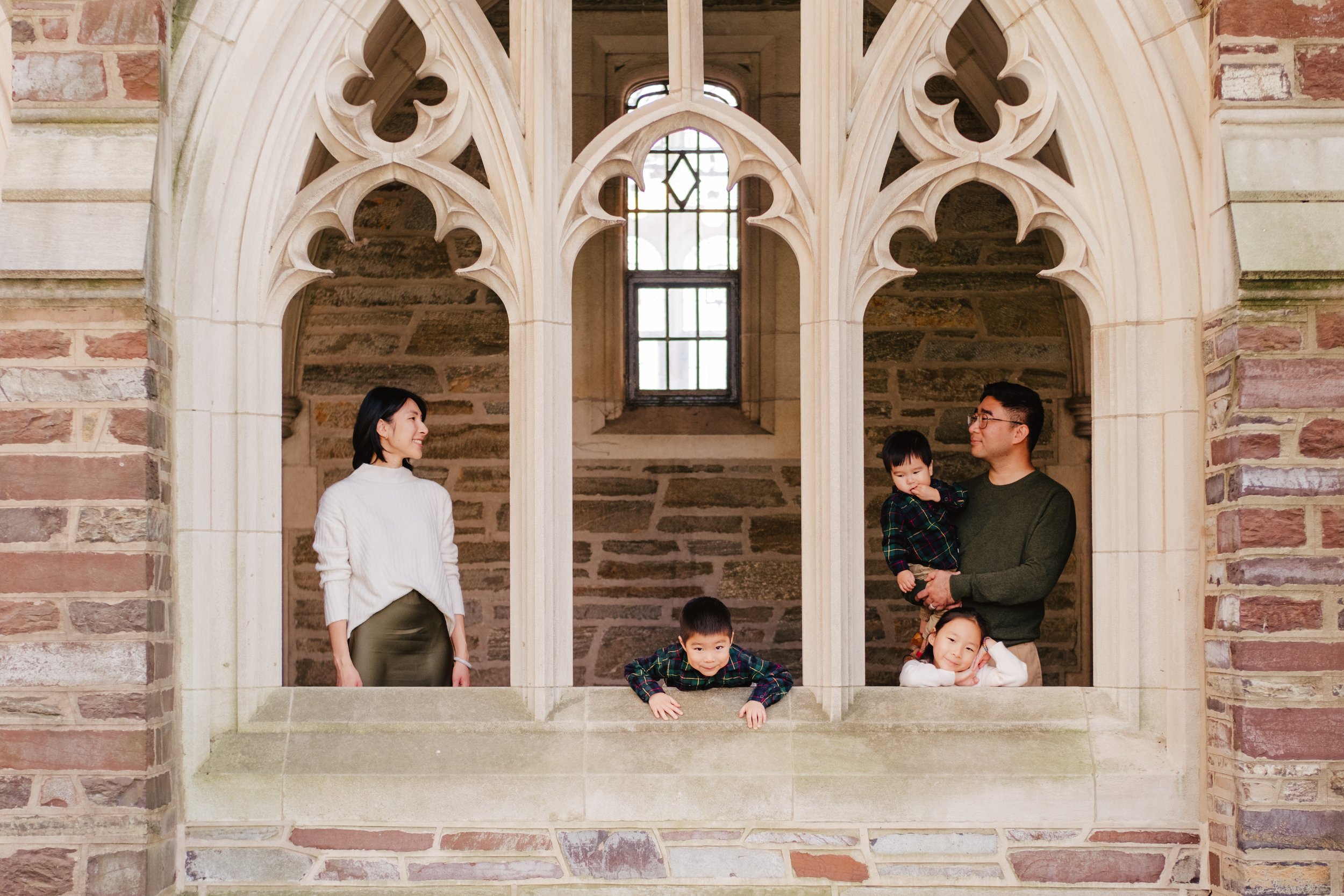 A family of four on a stone balcony with Gothic-style arches, inside a historic building with brick and stone walls, and a window in the background.