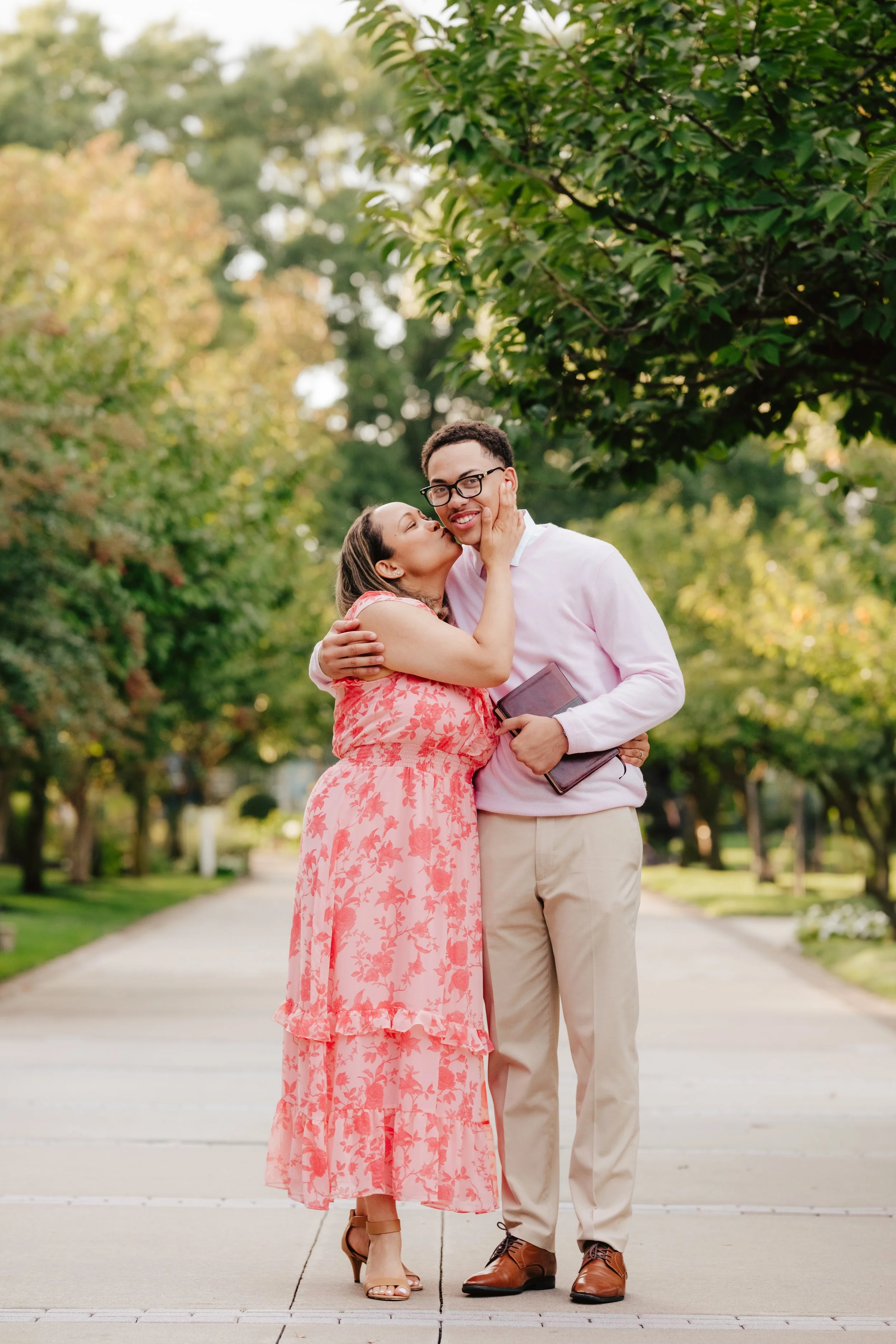 A woman in a pink floral dress kisses a man in a light pink sweater and beige pants on the cheek, standing on a park pathway lined with green trees.