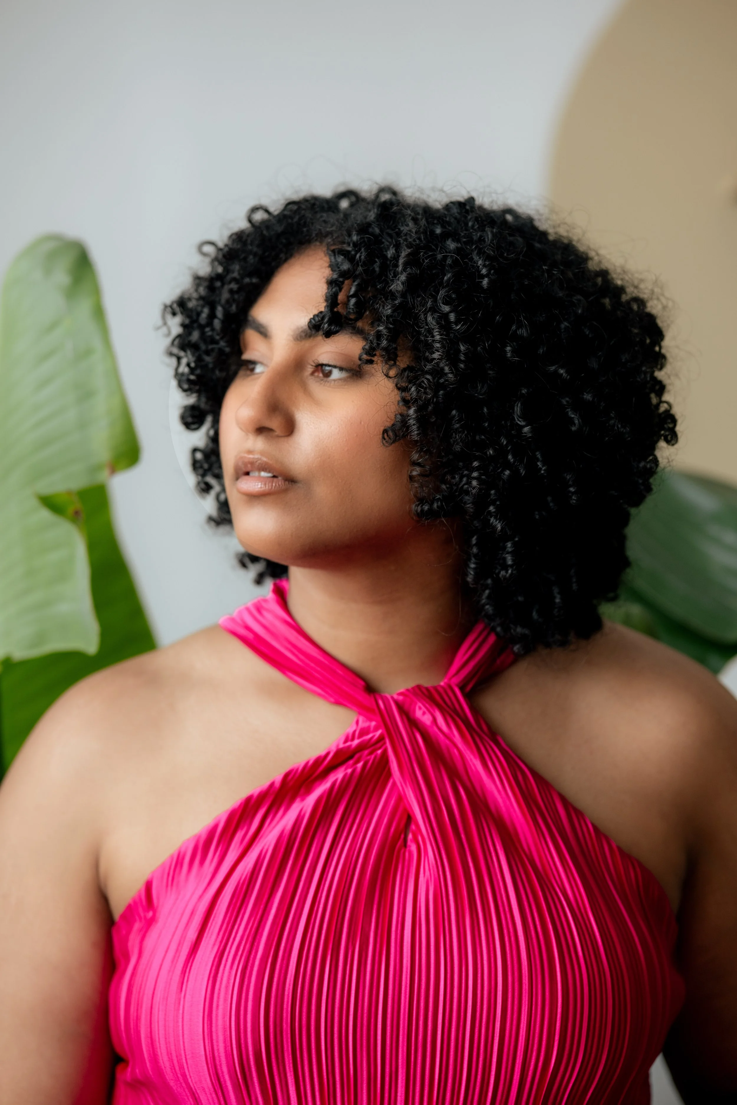A woman in a pink, pleated halter dress with curly black hair standing near large green tropical leaves.