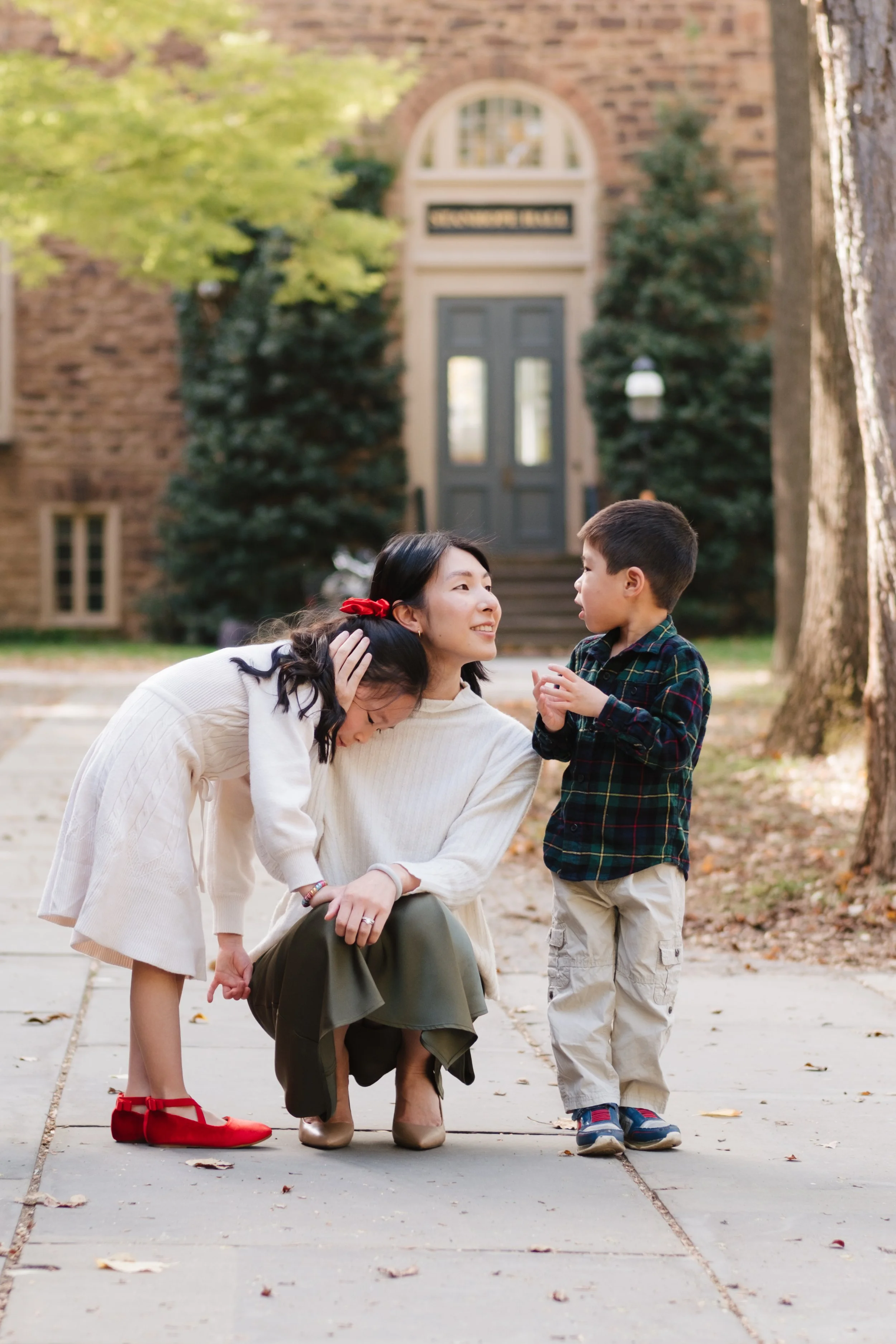 A woman kneeling on a sidewalk, surrounded by two children, a girl and a boy, in front of a brick house with greenery.