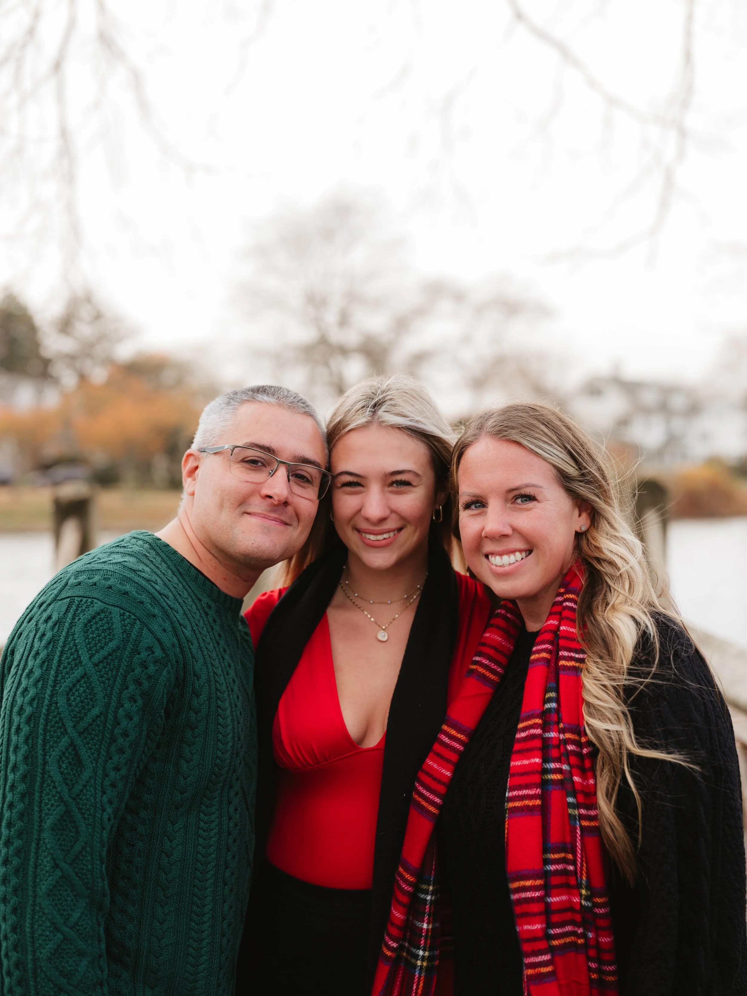 Three friends smiling outdoors on a cloudy day, standing close together near a body of water with trees and houses in the background.