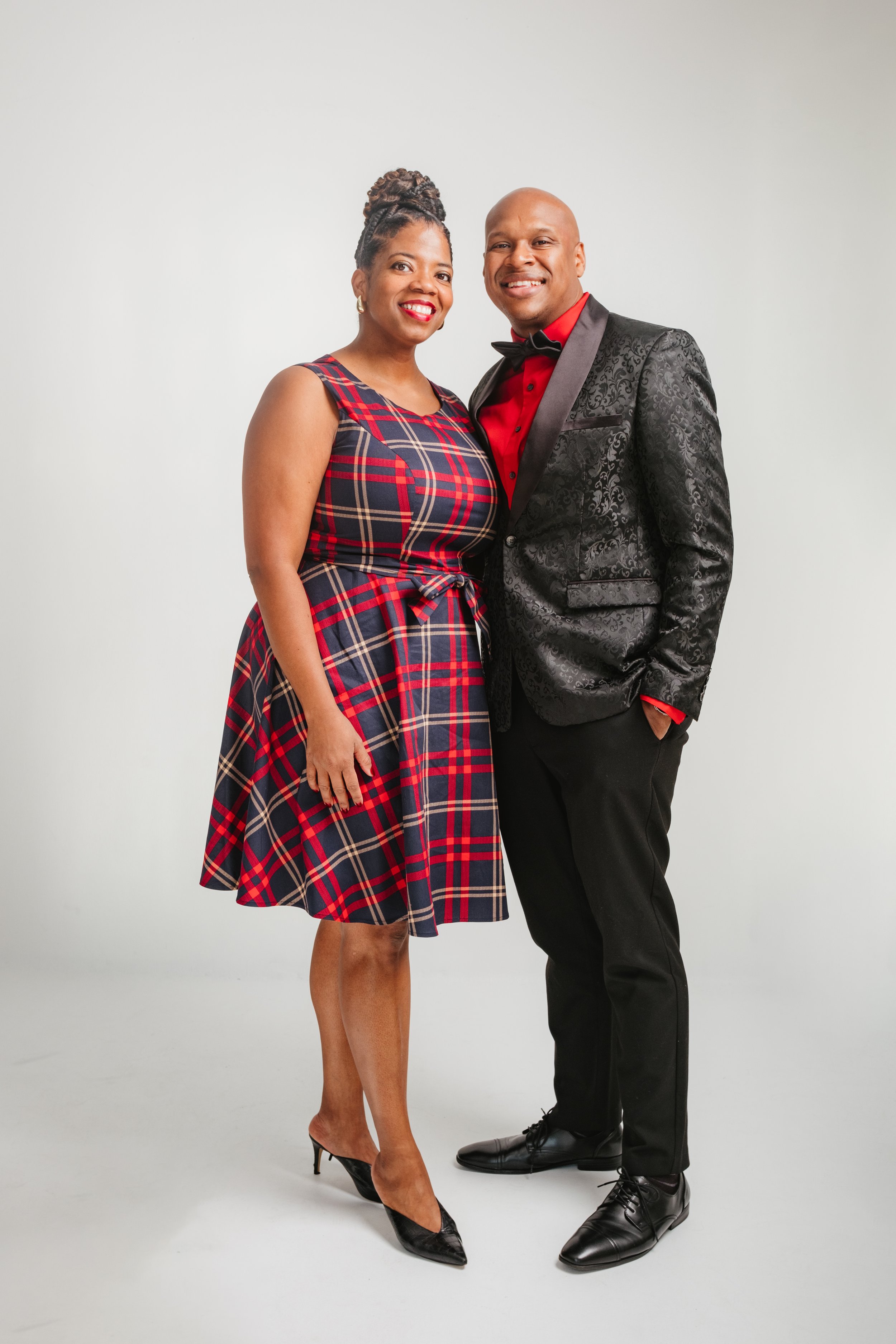 A man and woman dressed formally, standing close together and smiling at the camera, against a plain background.