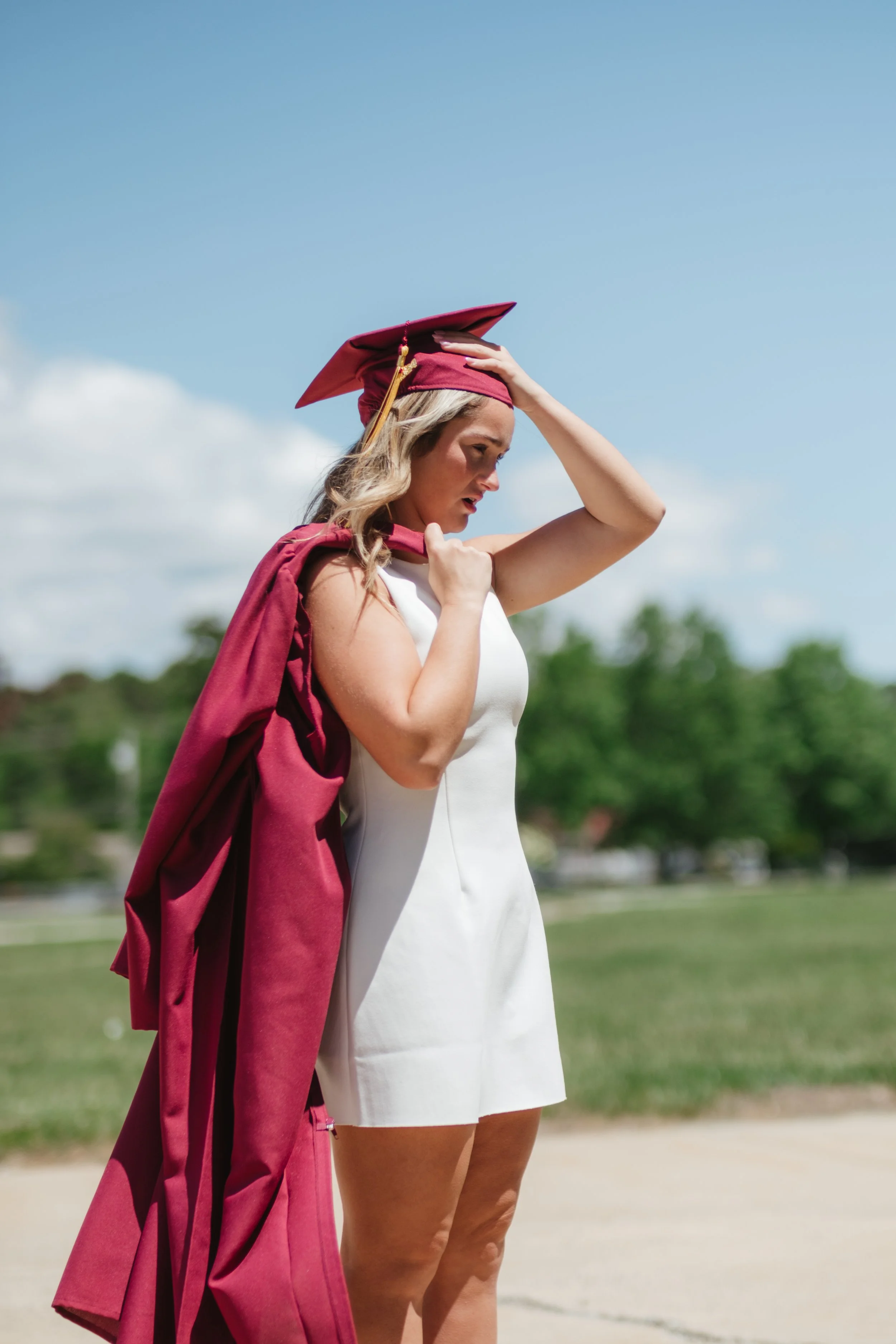 Young woman in a white dress with a maroon graduation cap and gown over her shoulder, standing outdoors on a sunny day with a blue sky and green trees in the background.