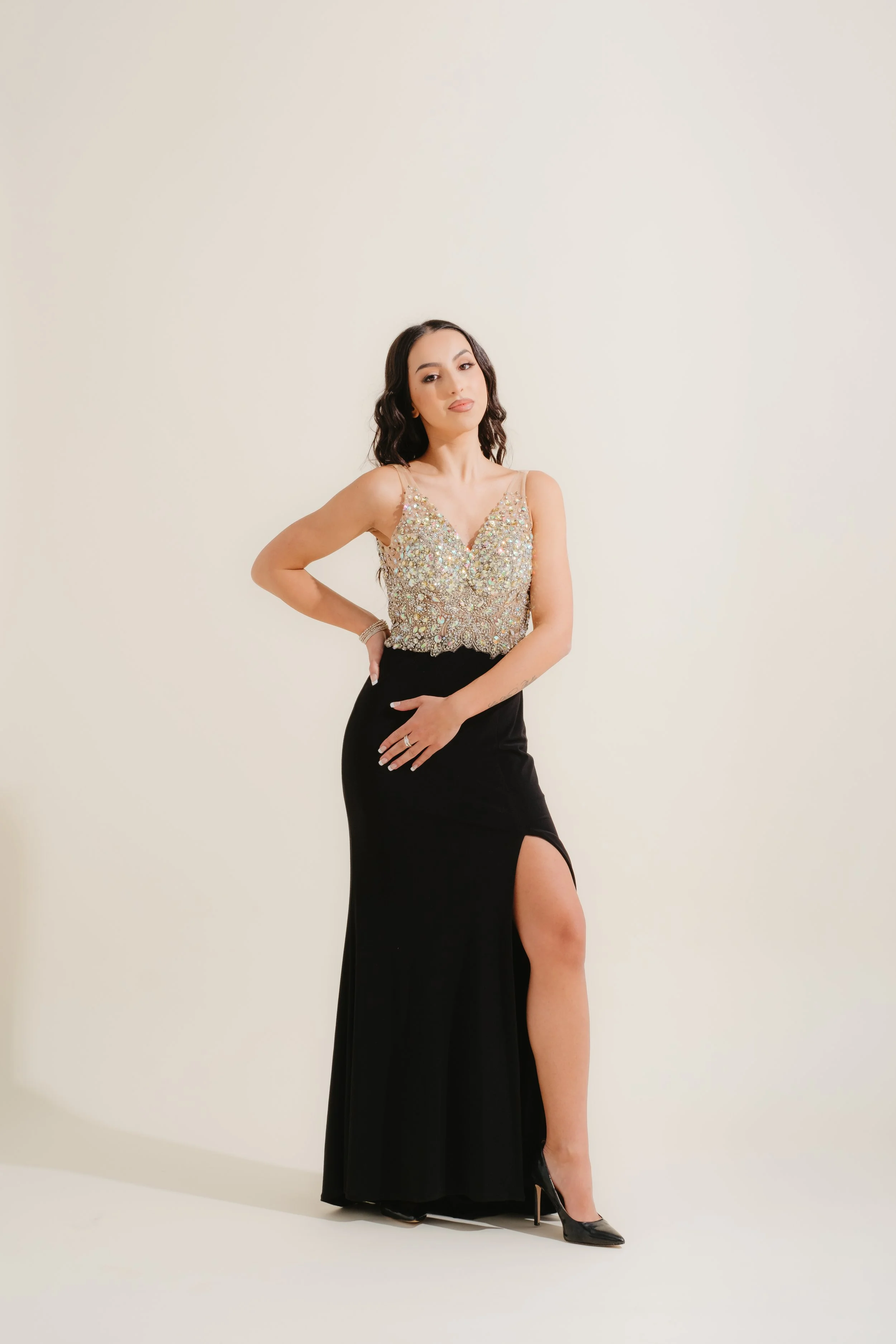 Woman in a sparkly sleeveless top and long black skirt with a high slit, standing against a plain light background