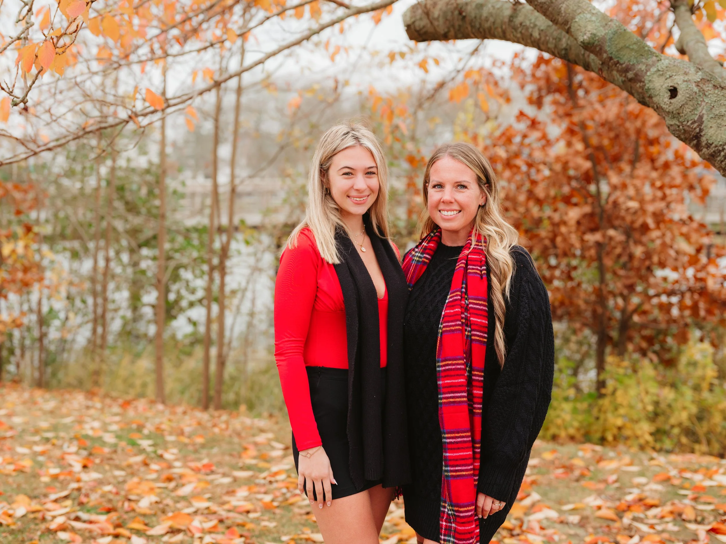 Two women standing outdoors in autumn with orange and brown leaves on trees and ground, smiling at the camera.