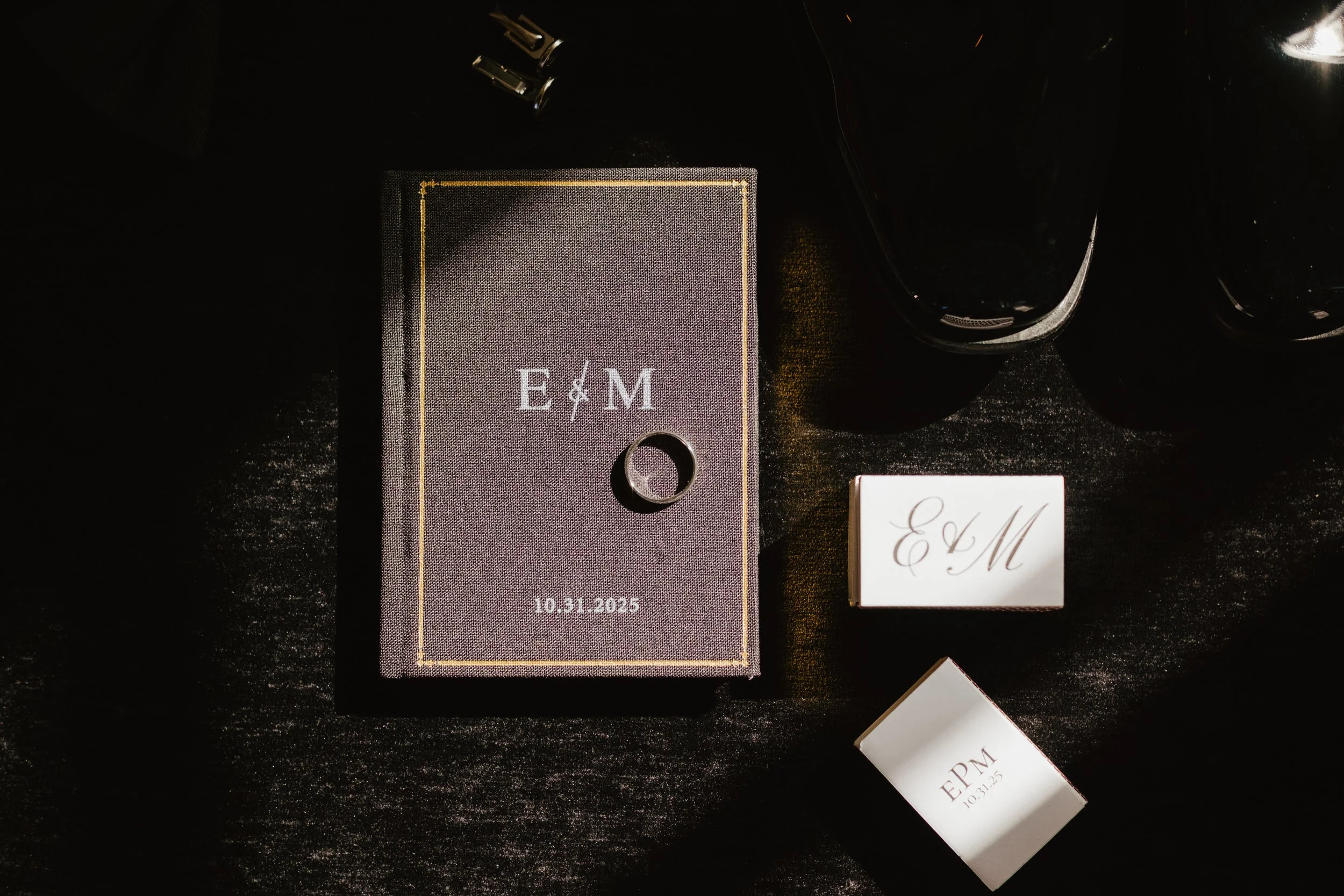 Invitation suite for a wedding with initials EM, date October 31, 2025, a black ring, and dark-colored sunglasses on a textured black surface.