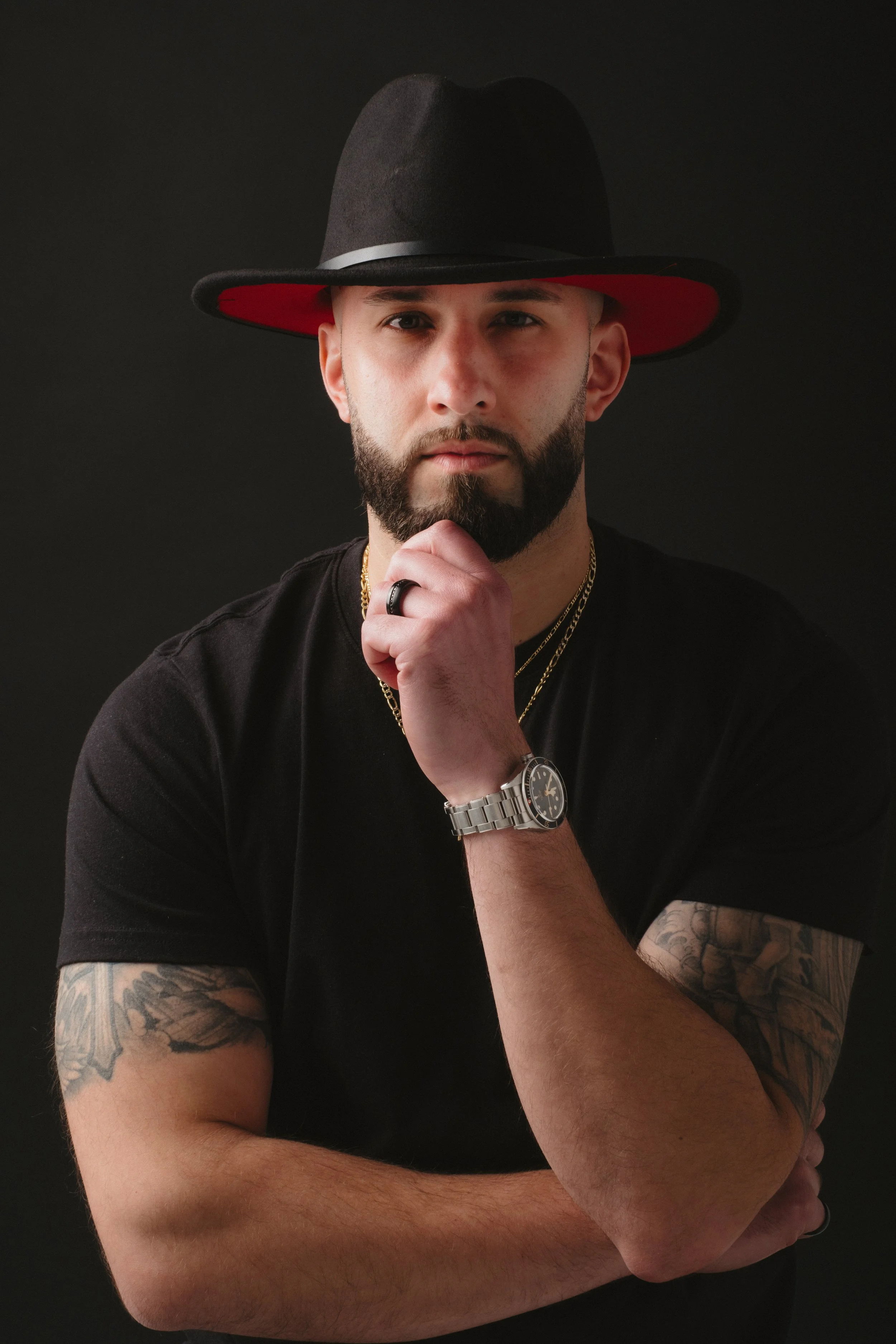 Portrait of a man with a beard wearing a black hat with a red underside, black T-shirt, gold chains, a wristwatch, a ring, and tattoos on his arms, standing against a dark background.