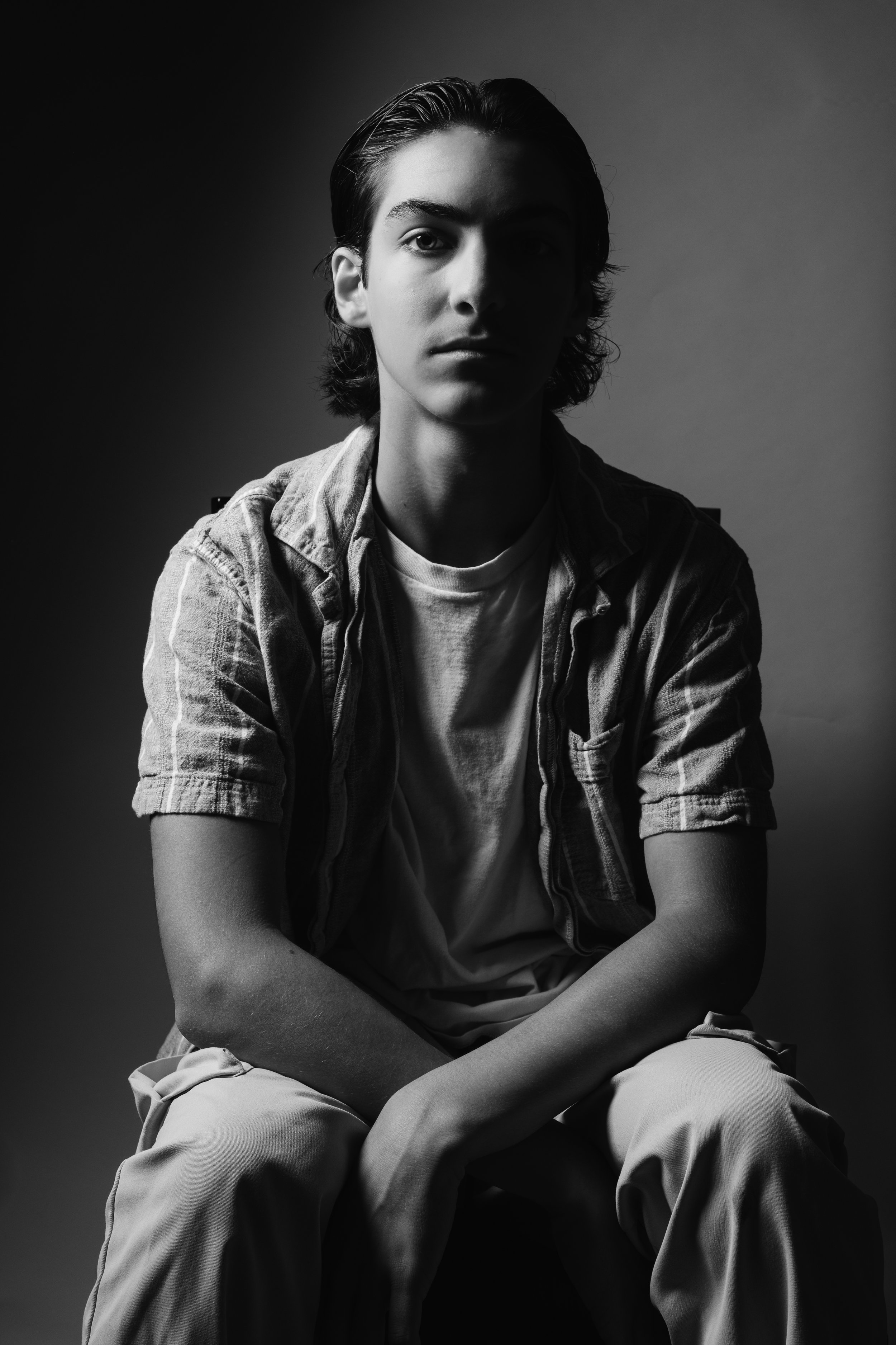 A young man with dark, slicked-back hair sitting against a plain background in black and white. He is wearing a short-sleeve shirt over a T-shirt and light-colored pants.