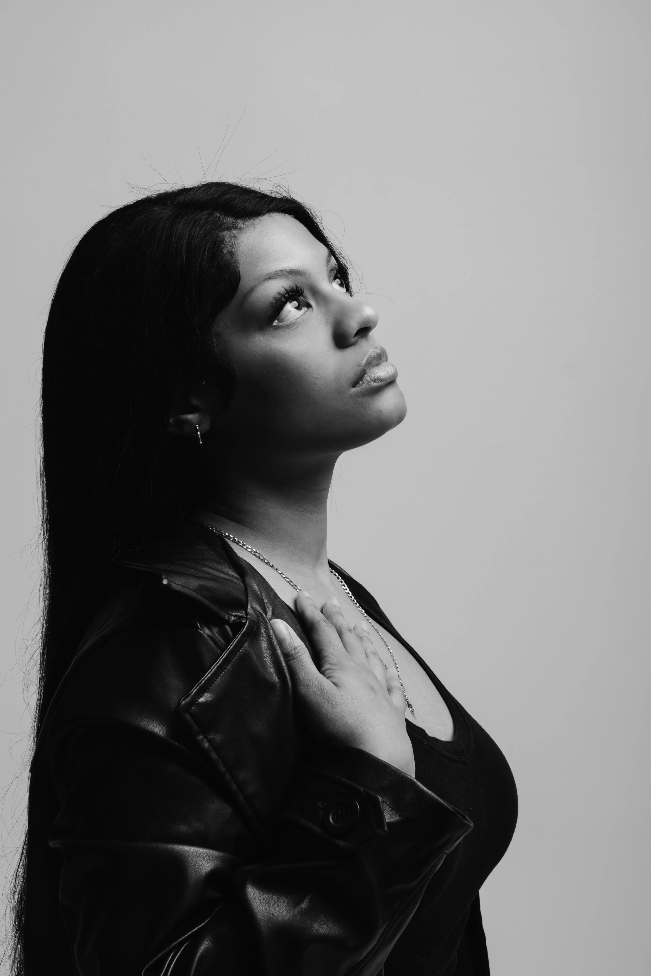Black and white portrait of a woman with long dark hair, wearing a leather jacket and a small necklace, looking upward with her hand on her chest, against a plain background.