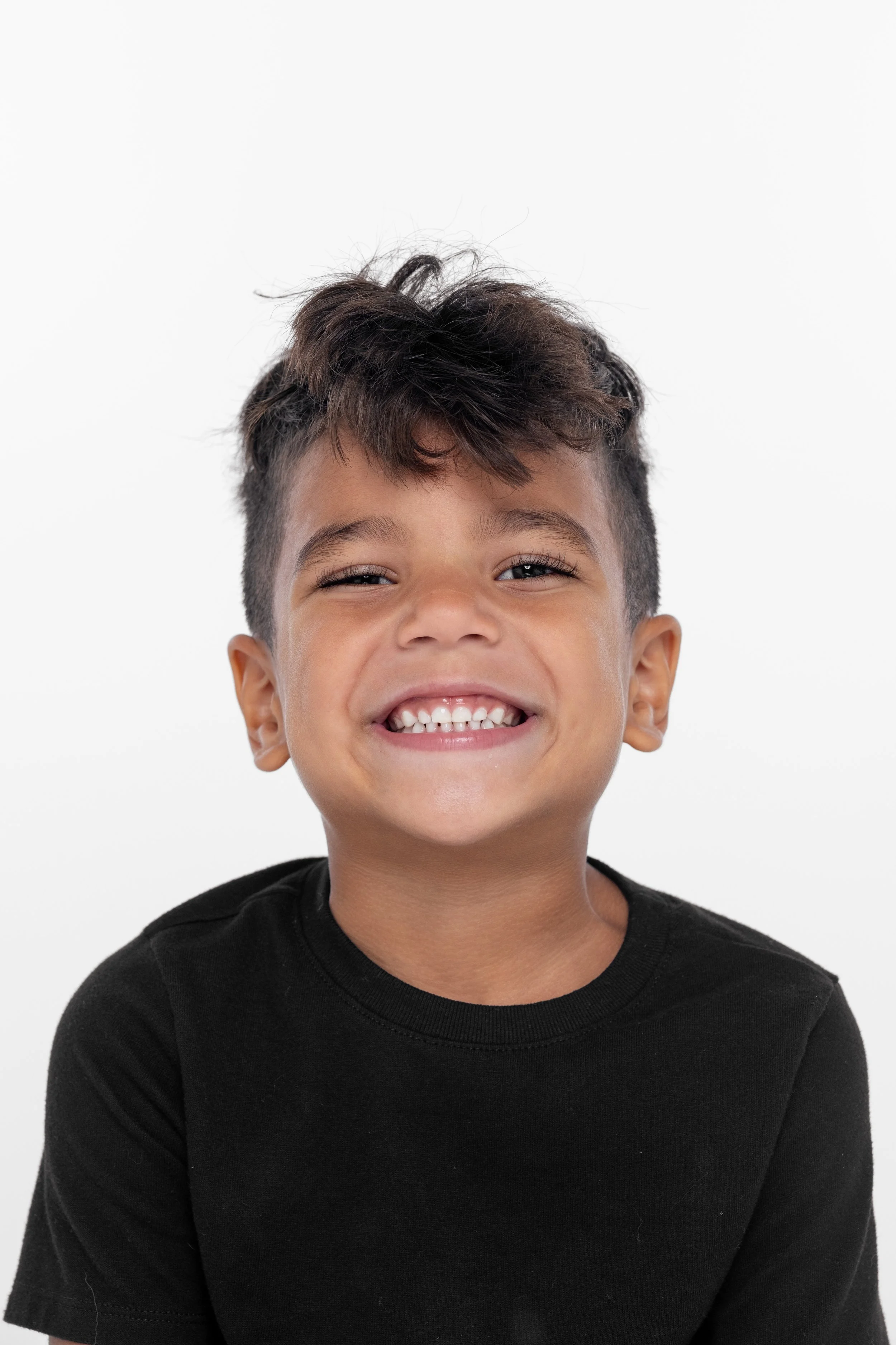 Close-up of a young boy with dark hair smiling broadly, showing his teeth, against a plain white background.