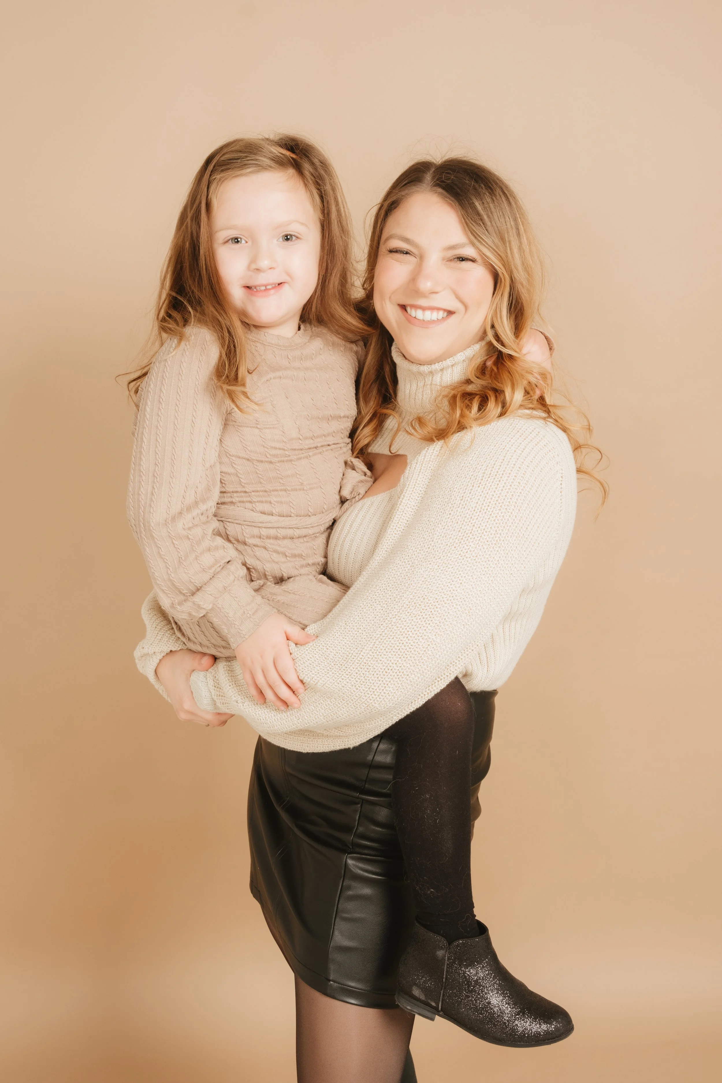 A woman with long, wavy, red hair smiling and holding a young girl with curly, red hair in her arms. Both are dressed in beige and cream-colored sweaters, and the girl is wearing black leggings and sparkly black boots. The background is a plain beige