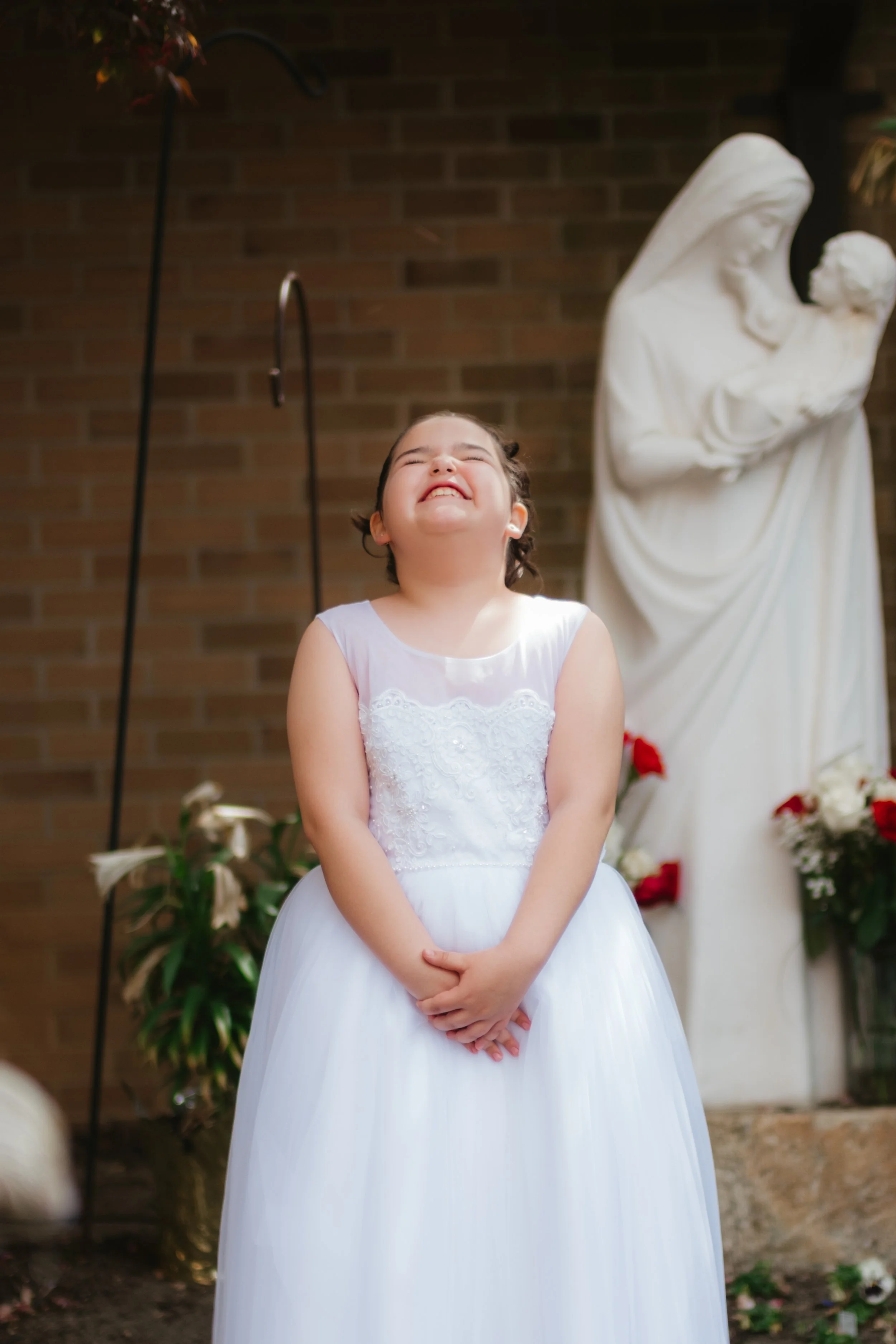 Young girl in a white dress smiling and laughing in front of a statue of the Virgin Mary holding baby Jesus, with a brick wall background.