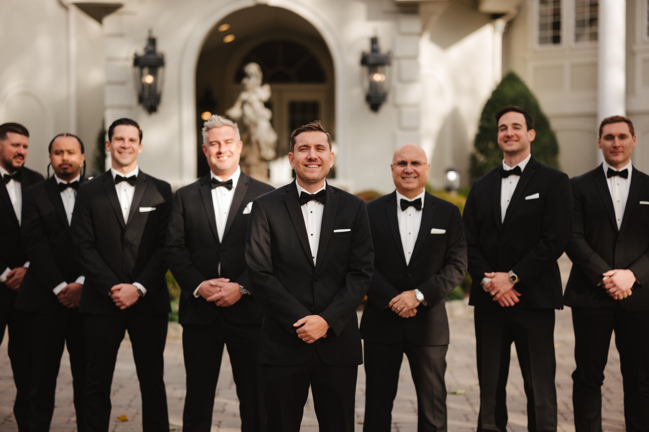 A group of men dressed in black tuxedos with bow ties, standing outdoors in front of a grand building, smiling at the camera.