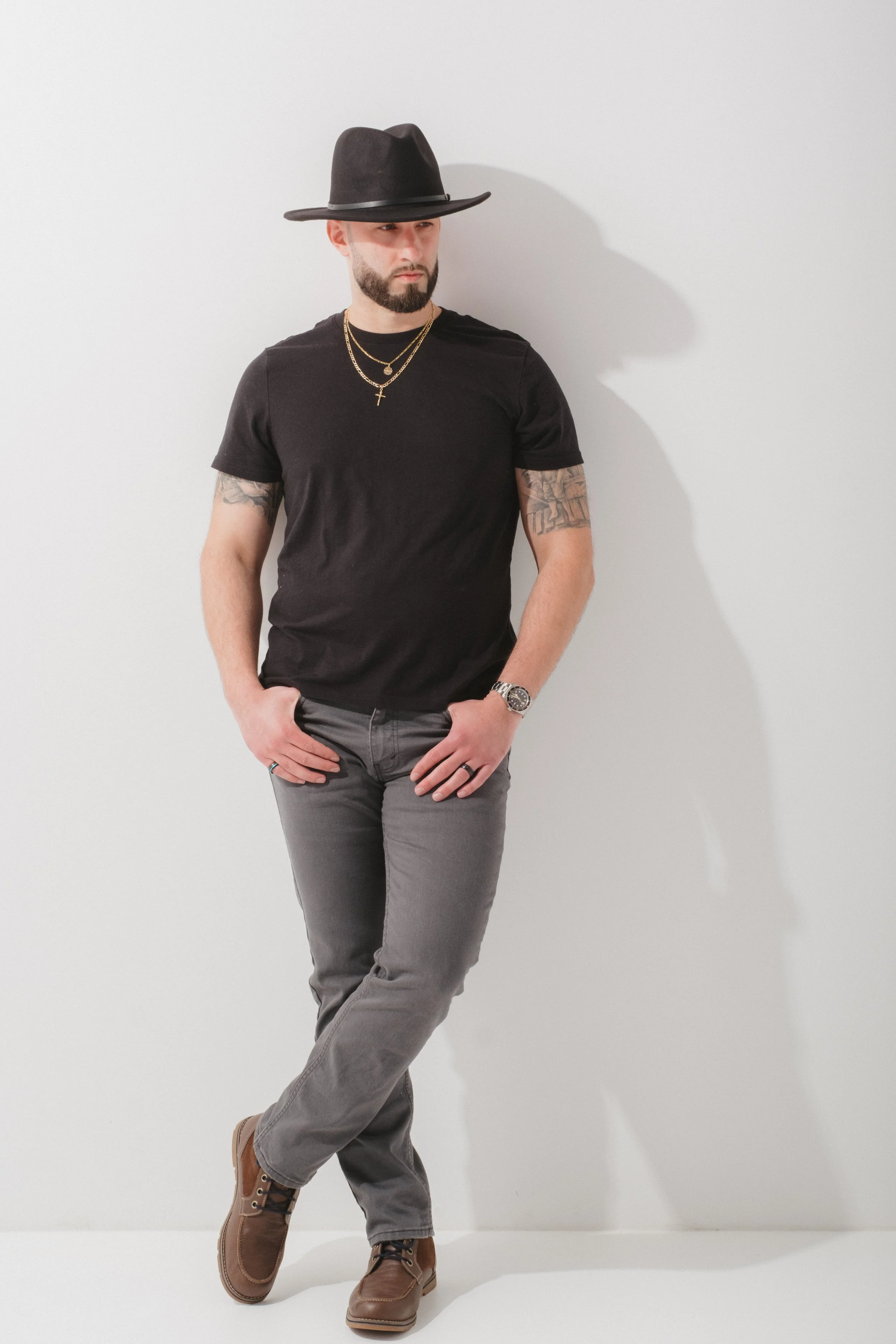 A man with a beard and tattoos, wearing a black t-shirt, gray jeans, brown shoes, a black wide-brim hat, gold chains, a watch, and rings, standing against a white background.