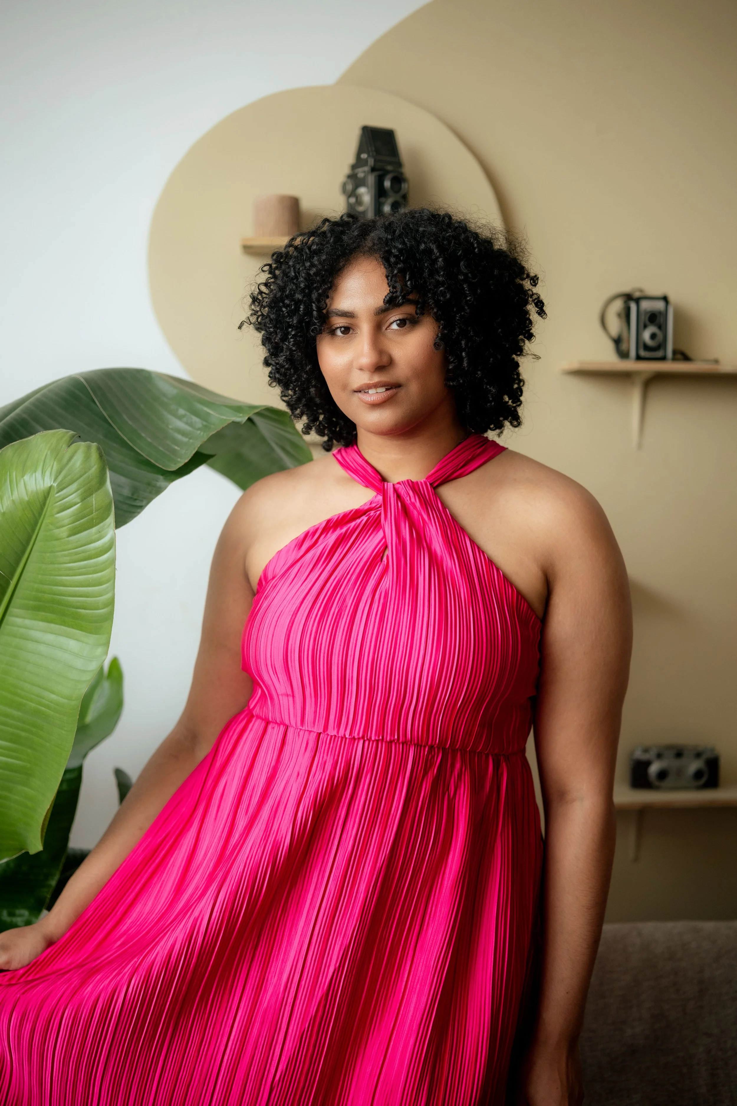 A woman with curly black hair wearing a bright pink pleated dress with a halter neck, standing indoors near a large green plant, with beige and white walls and shelves with stereo equipment in the background.