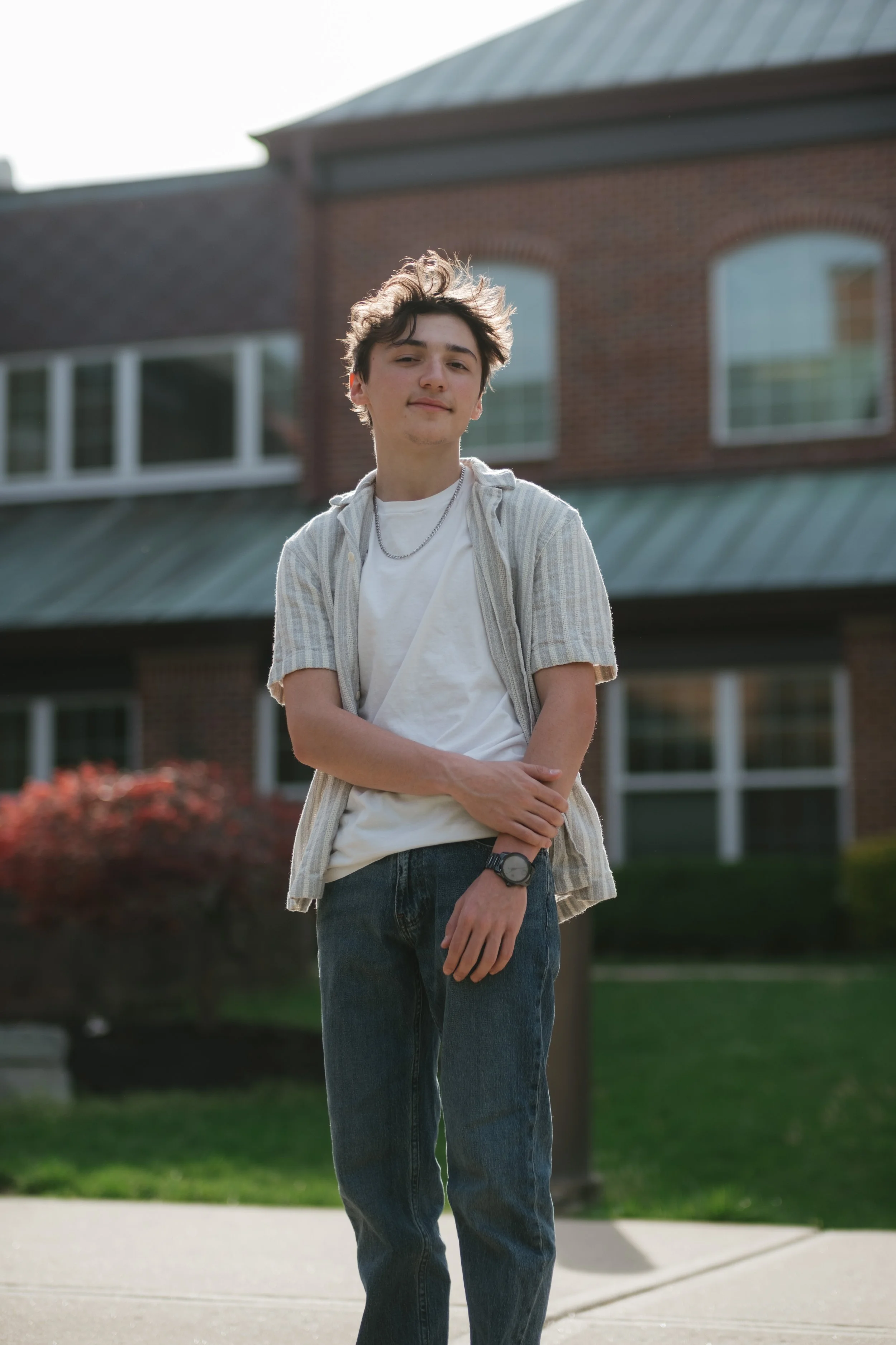 A young man with dark, tousled hair standing outdoors in front of a brick building, wearing a white T-shirt, a light-colored short-sleeve button-up shirt, jeans, a watch, and a necklace, with one arm crossed and the other resting on his stomach.