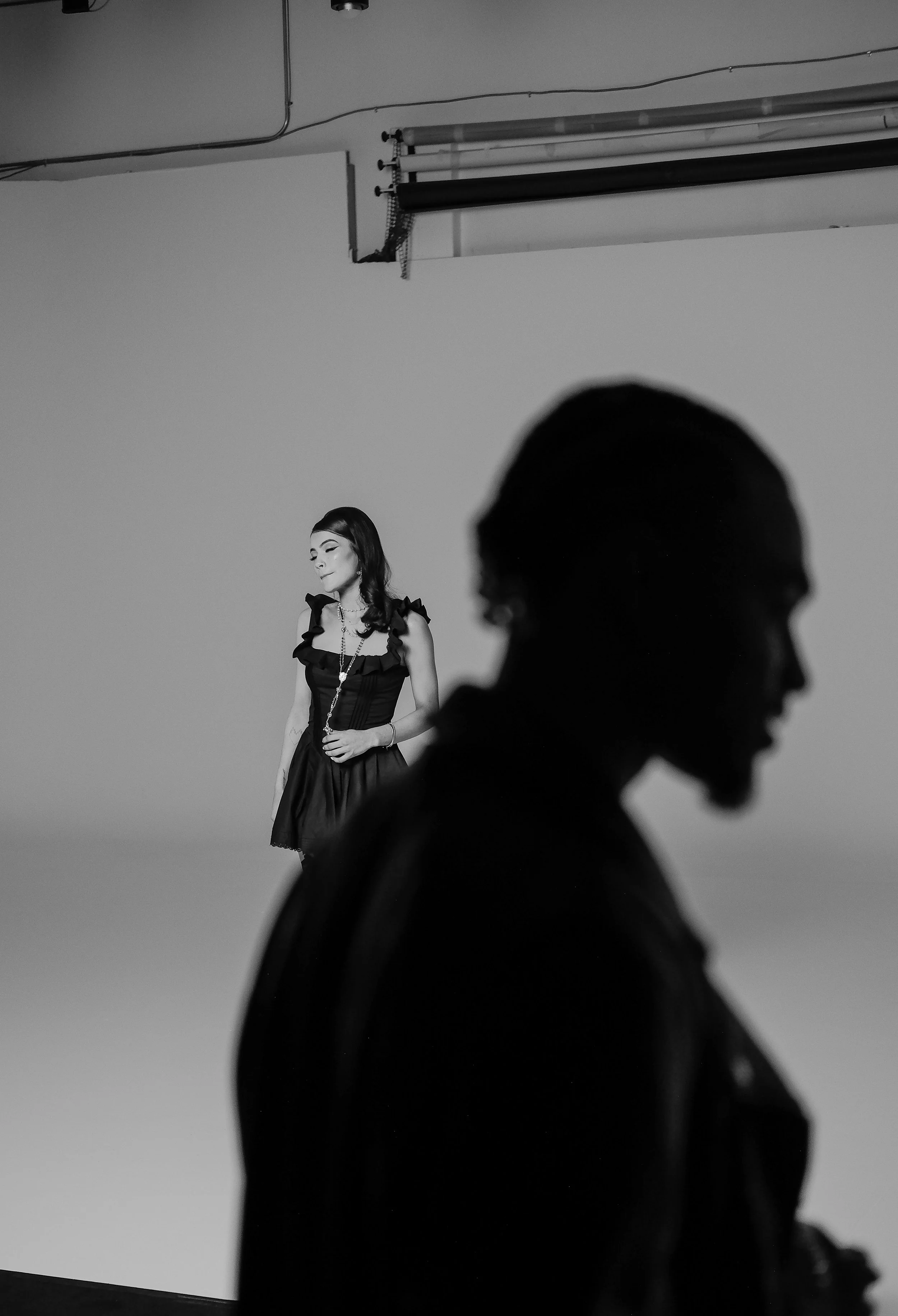 A black and white photo of a woman standing in the background with her eyes closed, and a silhouette of a man in the foreground, both indoors.