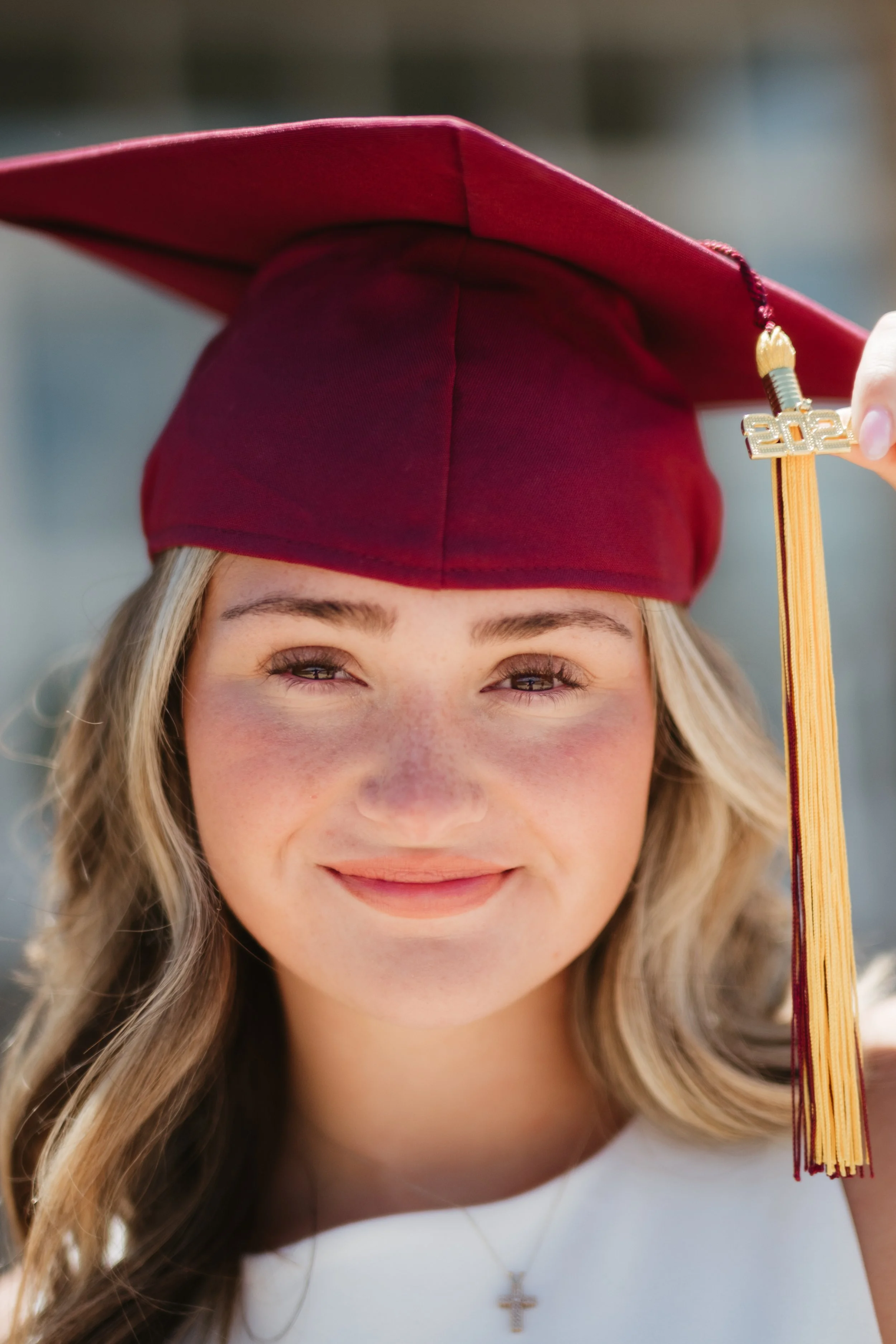 Close-up of a young woman wearing a maroon graduation cap, smiling, with a small cross necklace visible, holding a tassel with '2022' attached.