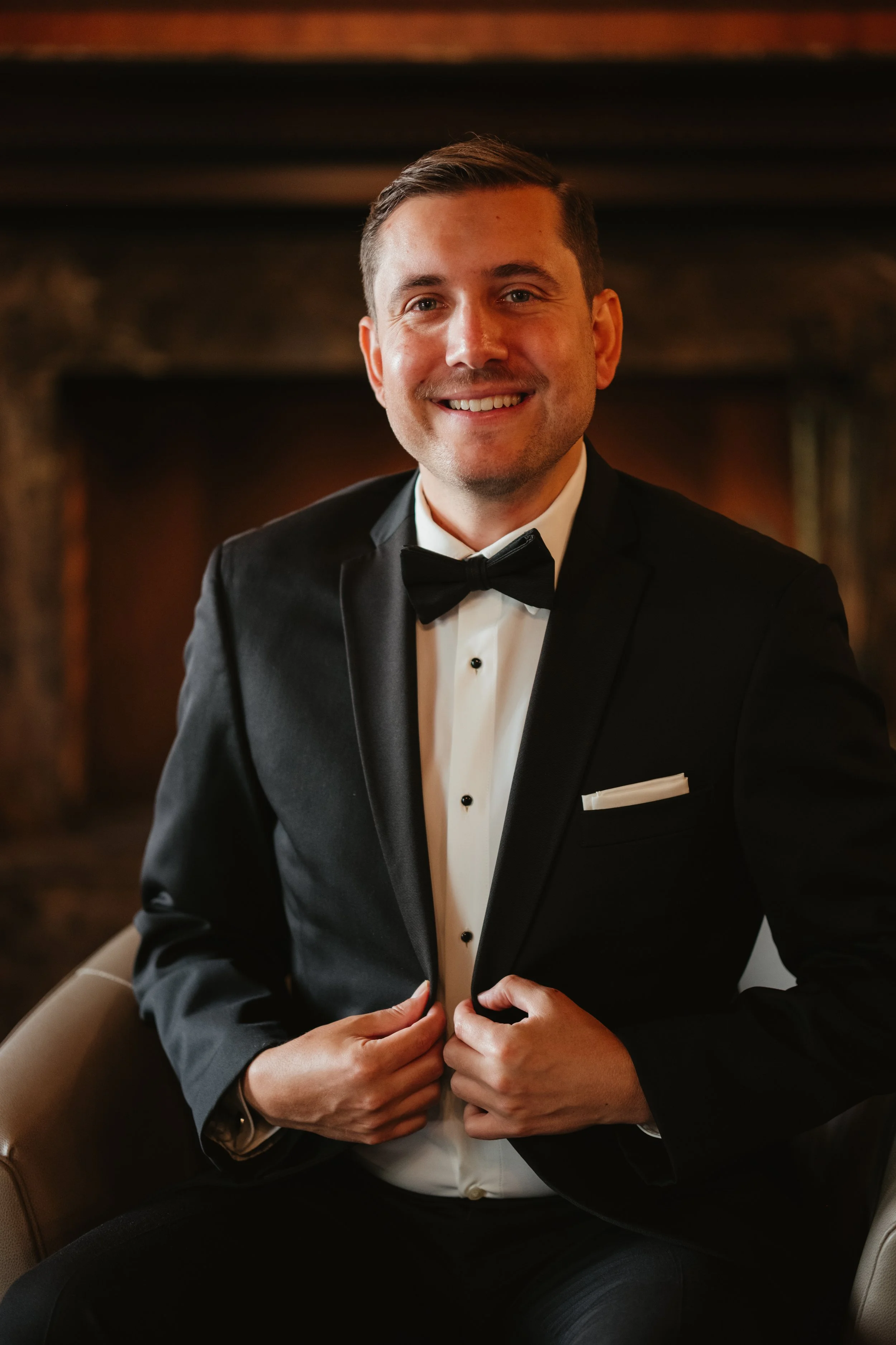 A man in a tuxedo, smiling, adjusting his jacket, sitting in a warmly lit room.