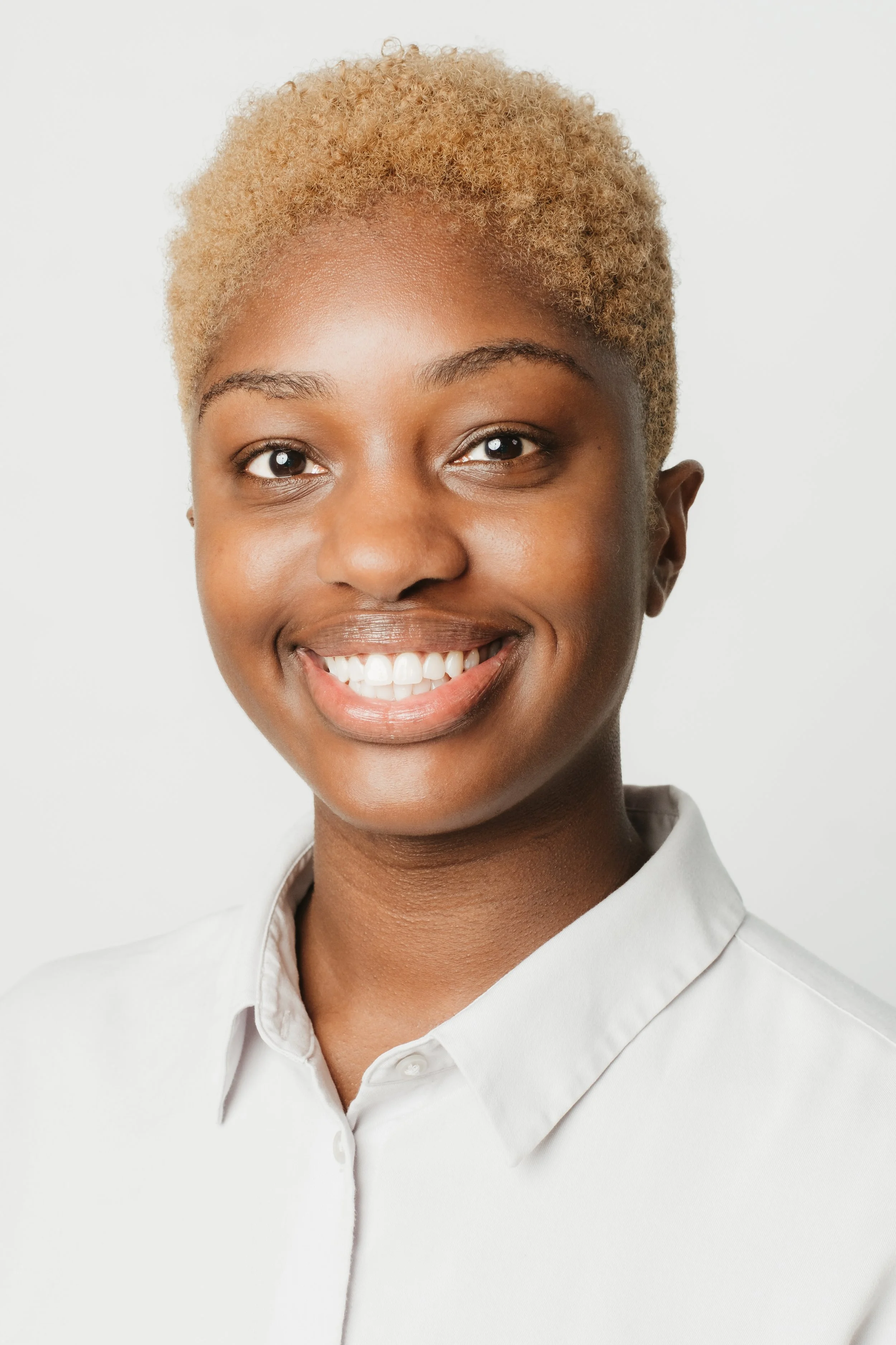 Close-up of a smiling young woman with short blond curly hair, wearing a white collared shirt, against a plain white background.