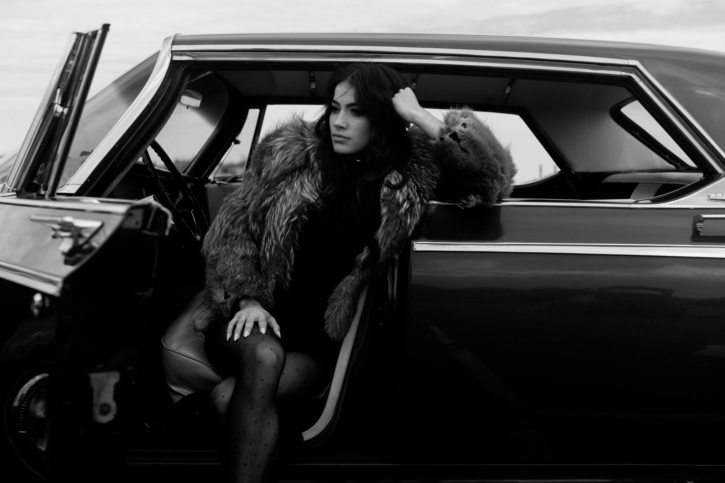 A woman with dark hair and a fur coat sitting inside a classic car with the door open, looking away thoughtfully.