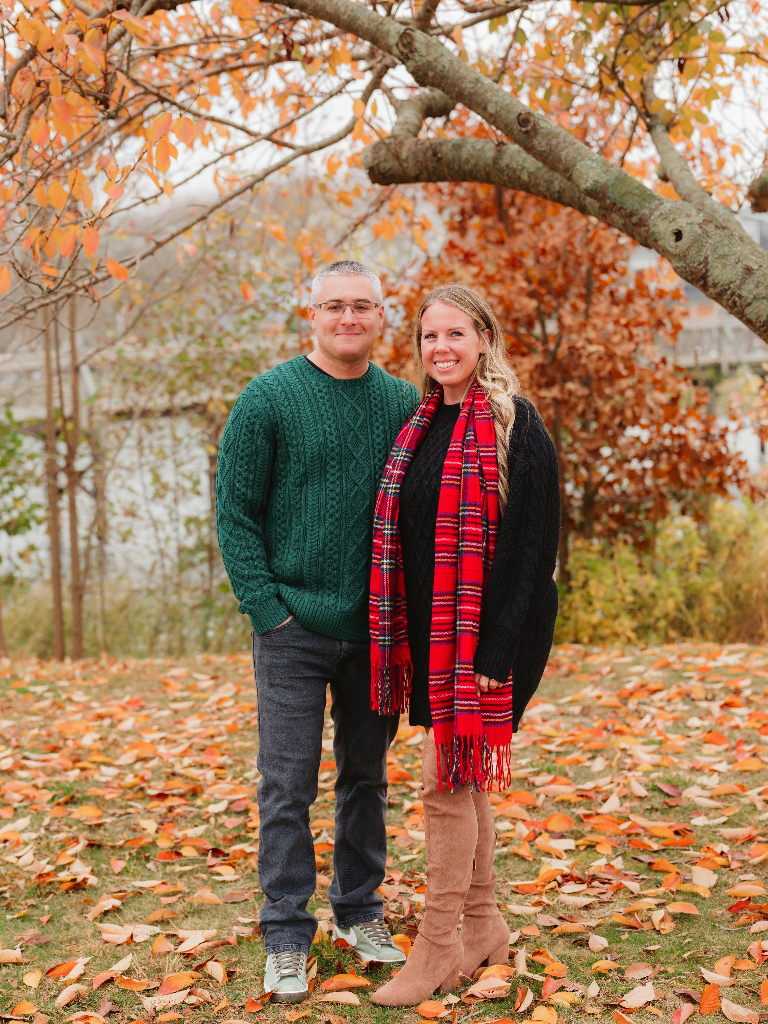 A man and a woman standing outdoors under a tree with orange and red autumn leaves, smiling at the camera, wearing fall clothing, with fallen leaves on the ground.