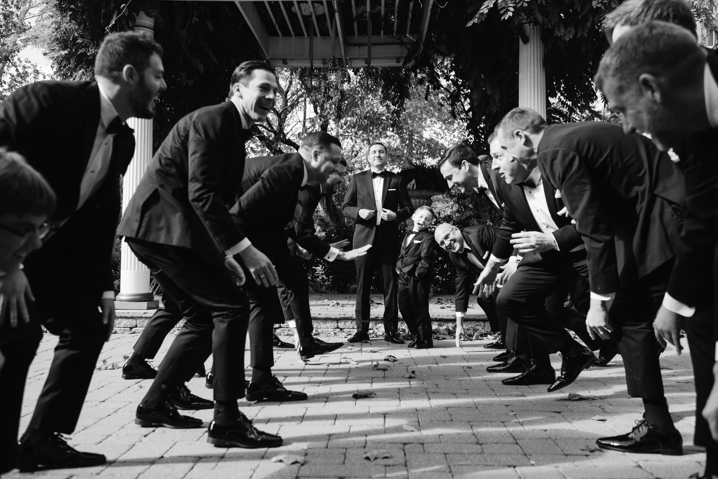 Black and white photo of a group of men in tuxedos participating in a traditional wedding dance, with a few children in tuxedos watching in the background. They are outdoors on a brick patio, smiling and bent forward in a playful manner.