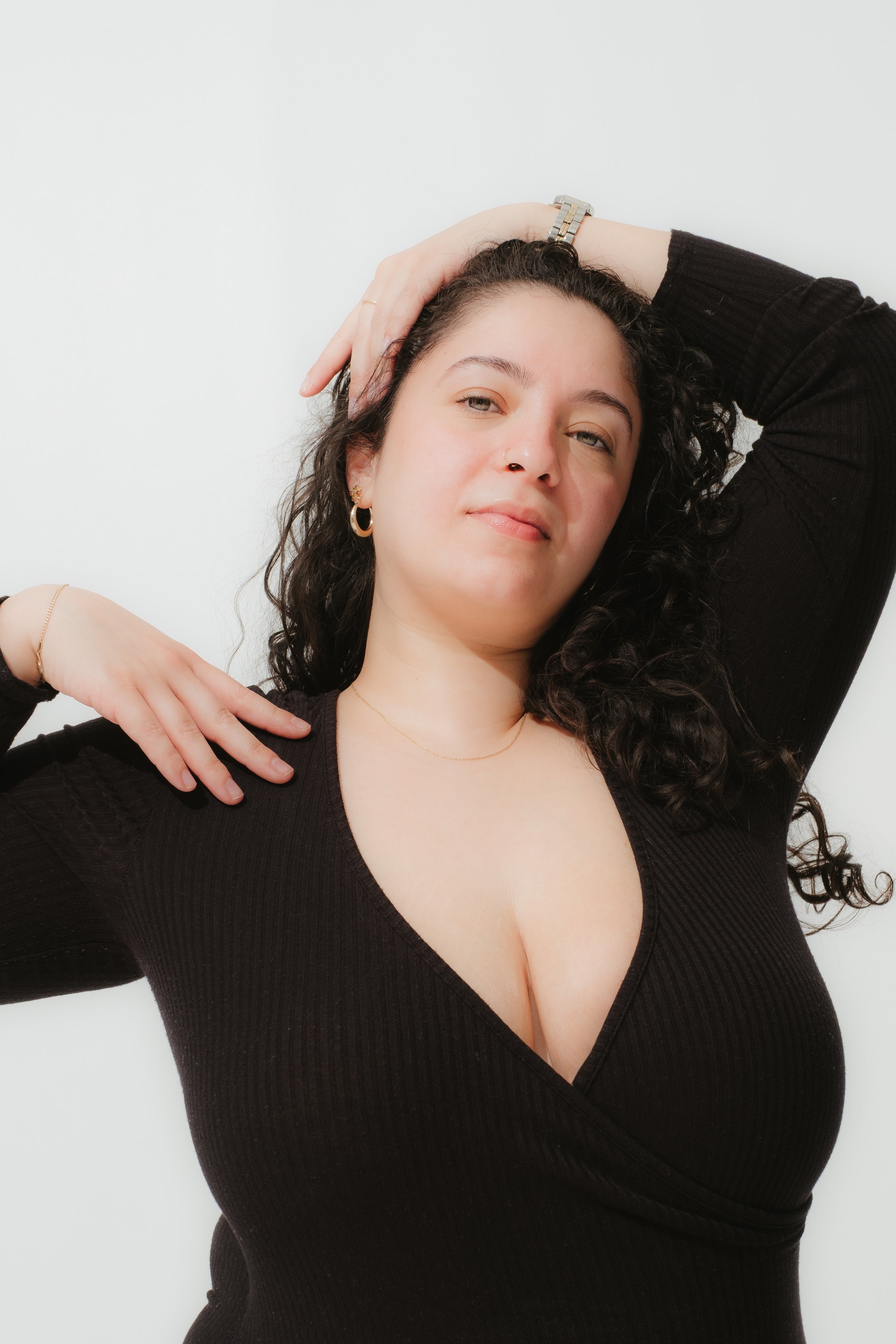 A woman with curly dark hair, wearing a black V-neck top, posing with one hand on her head and the other on her shoulder against a white background.