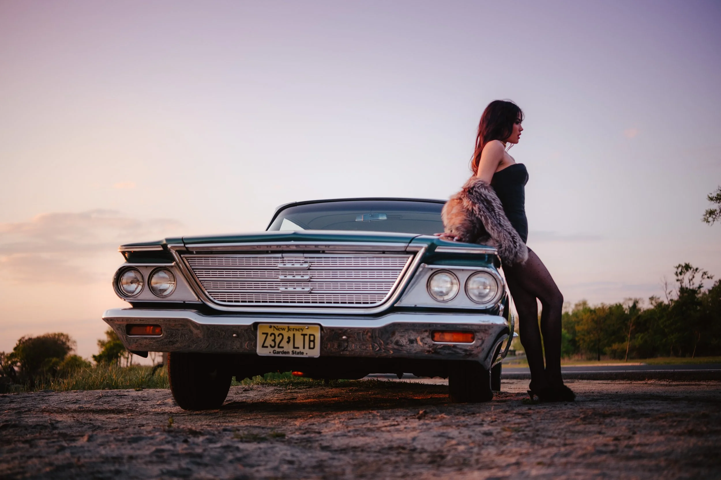 A woman sitting on the hood of a vintage black Chevrolet car with New Jersey license plate during sunset.