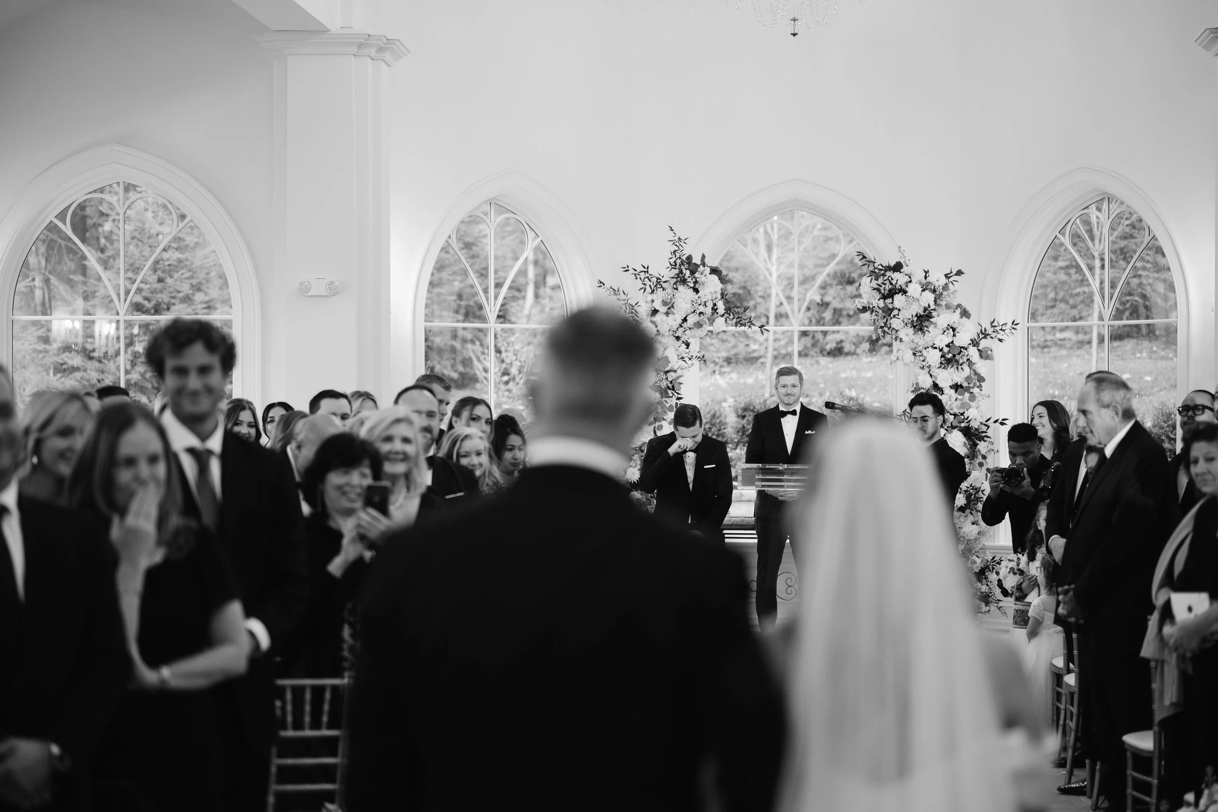 Black and white photo of a wedding ceremony inside a bright room with large windows. The bride and groom face each other in the foreground, with the officiant and wedding party behind them. Guests are seated and standing around the couple, some smili