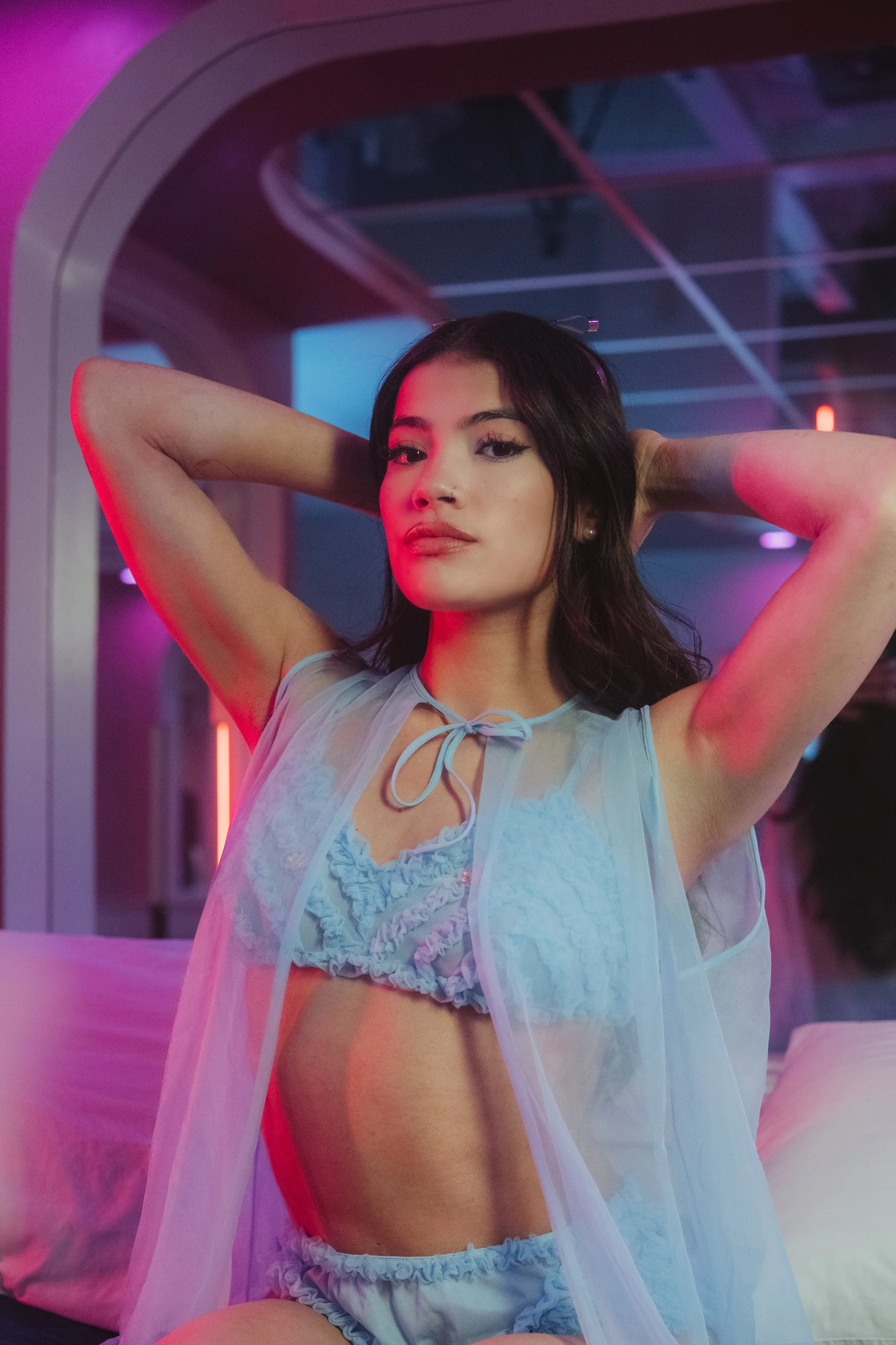 Young woman in lingerie posing in a neon-lit room with pink and blue lighting.