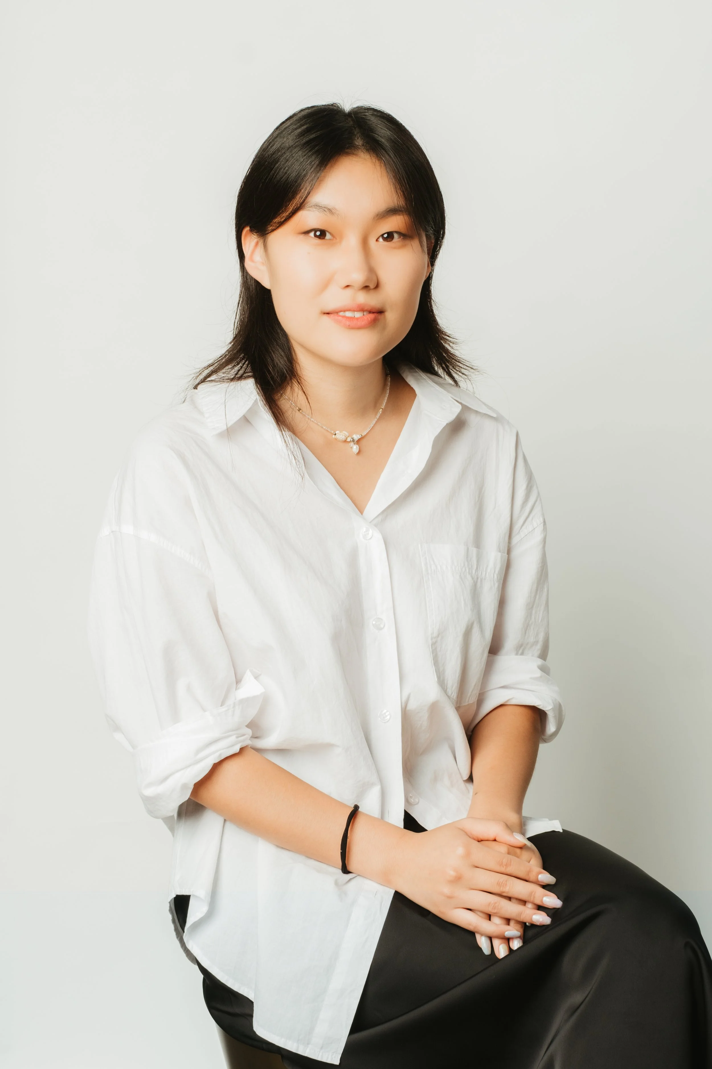 A young woman with black hair, wearing a white shirt and black skirt, sitting against a plain white background.
