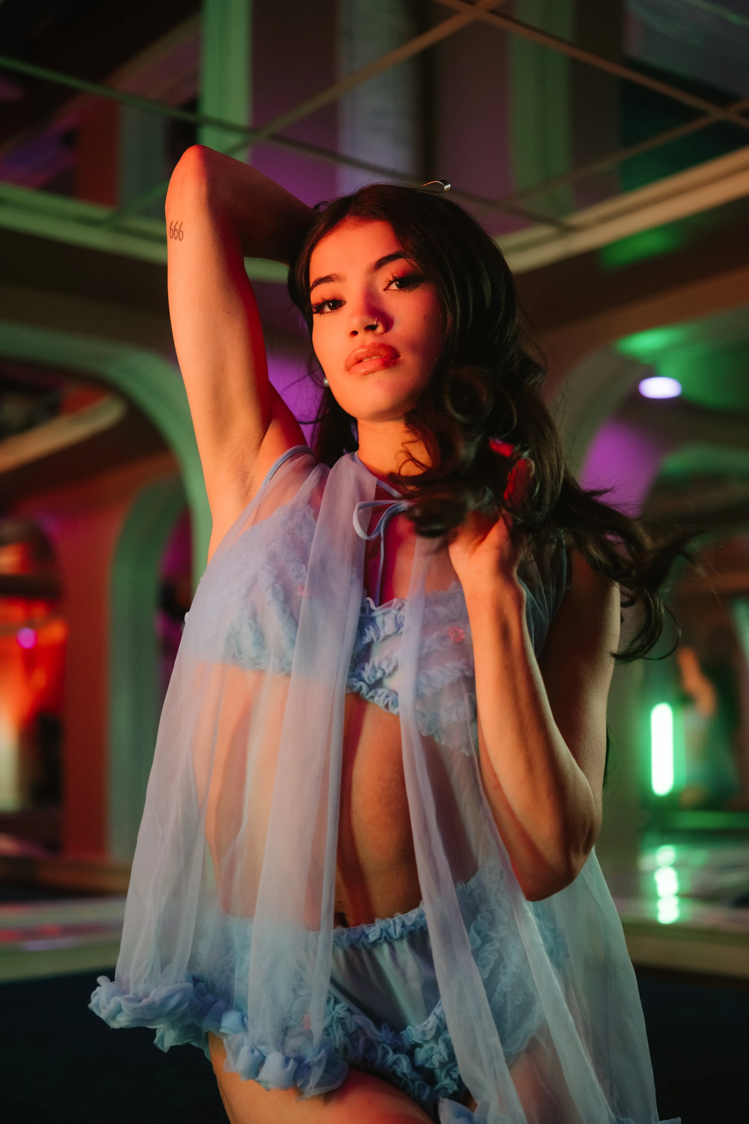 A woman in sheer lingerie posing with one arm raised behind her head in an indoor setting with colorful lighting.