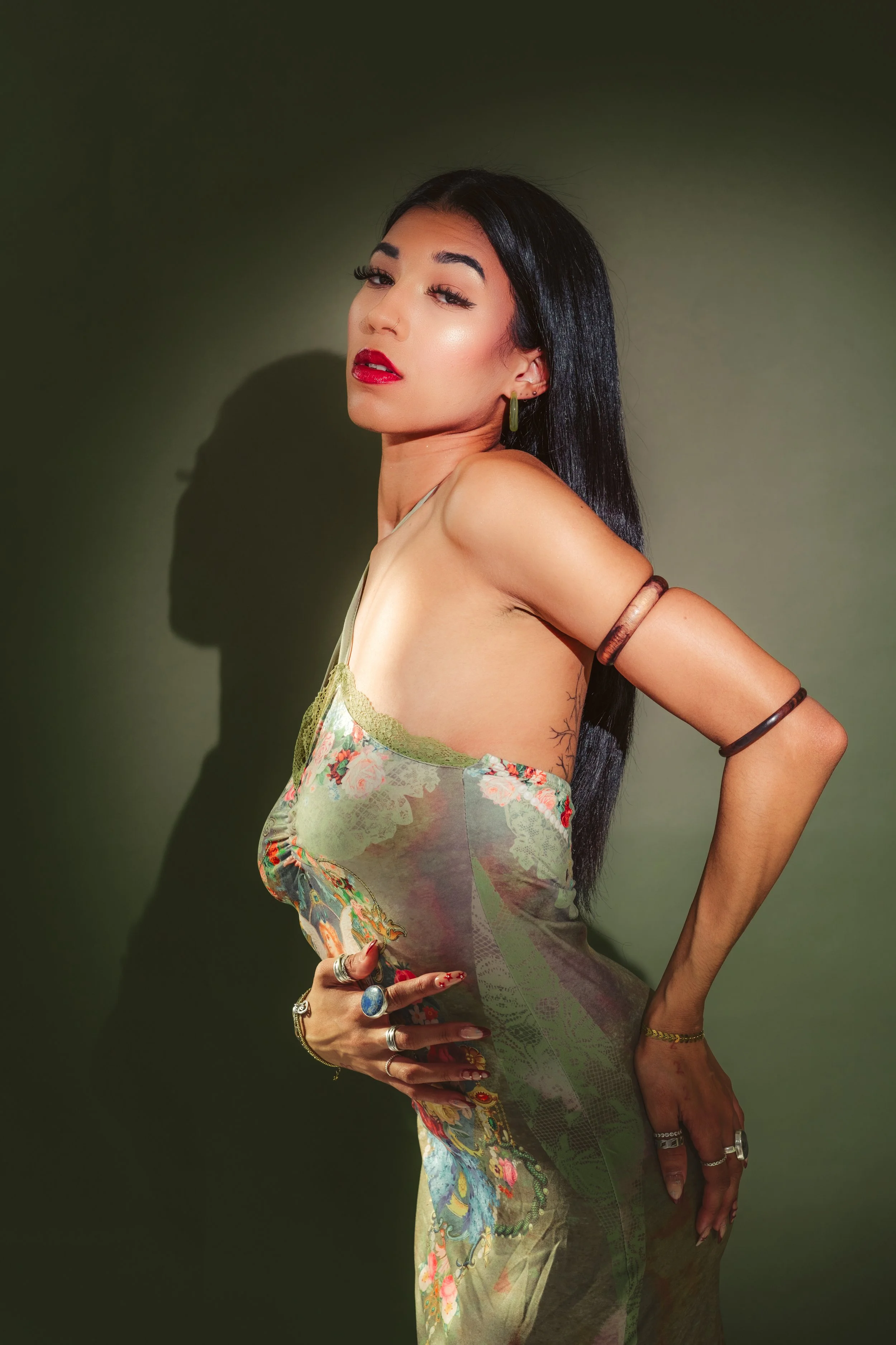 A woman with long black hair poses against a dark green background, wearing a floral dress with spaghetti straps. She has red lipstick, earrings, multiple rings, and bracelets, with her hand on her hip, and is looking at the camera.