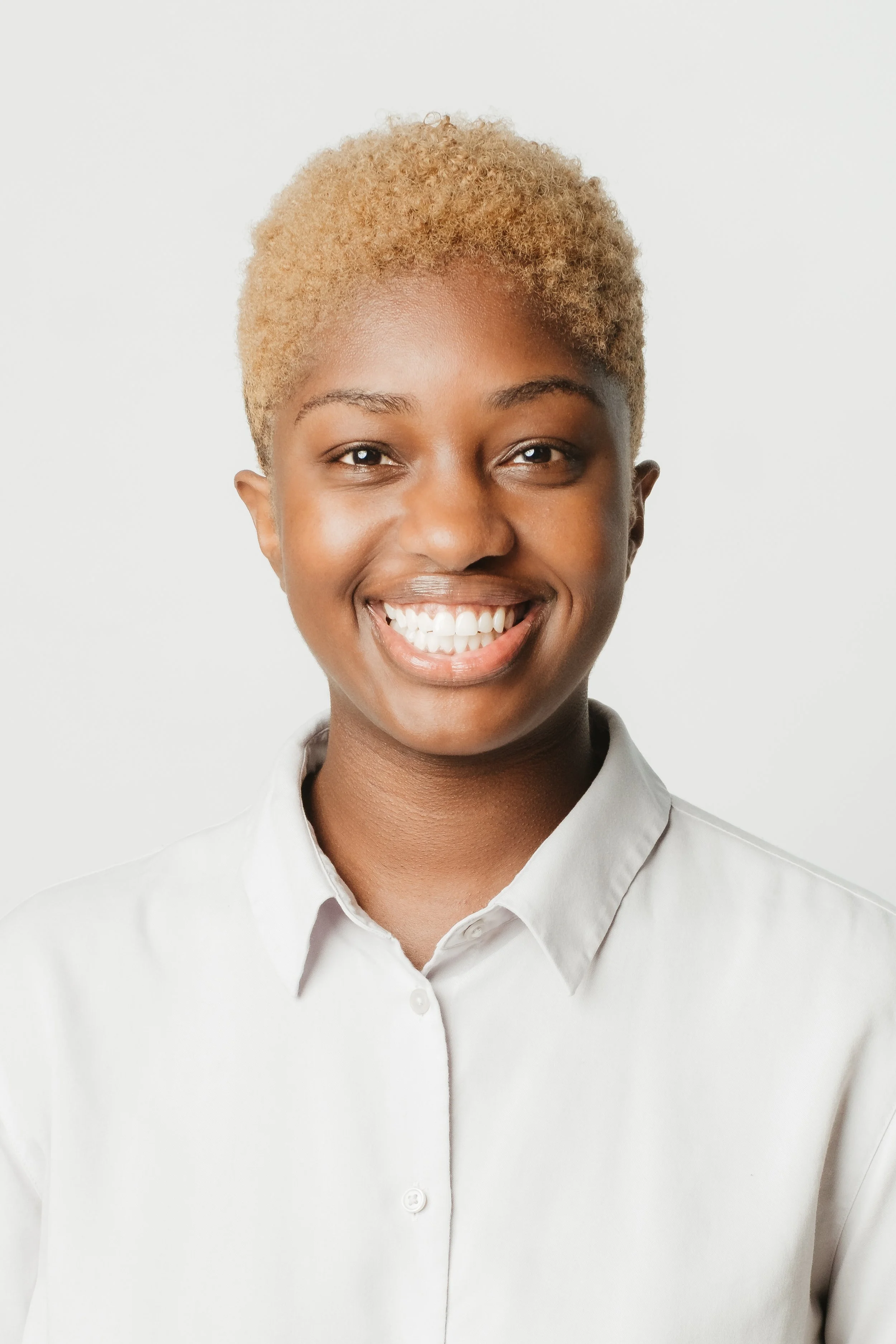 A young woman with short, curly blonde hair smiling and wearing a white button-up shirt against a plain white background.