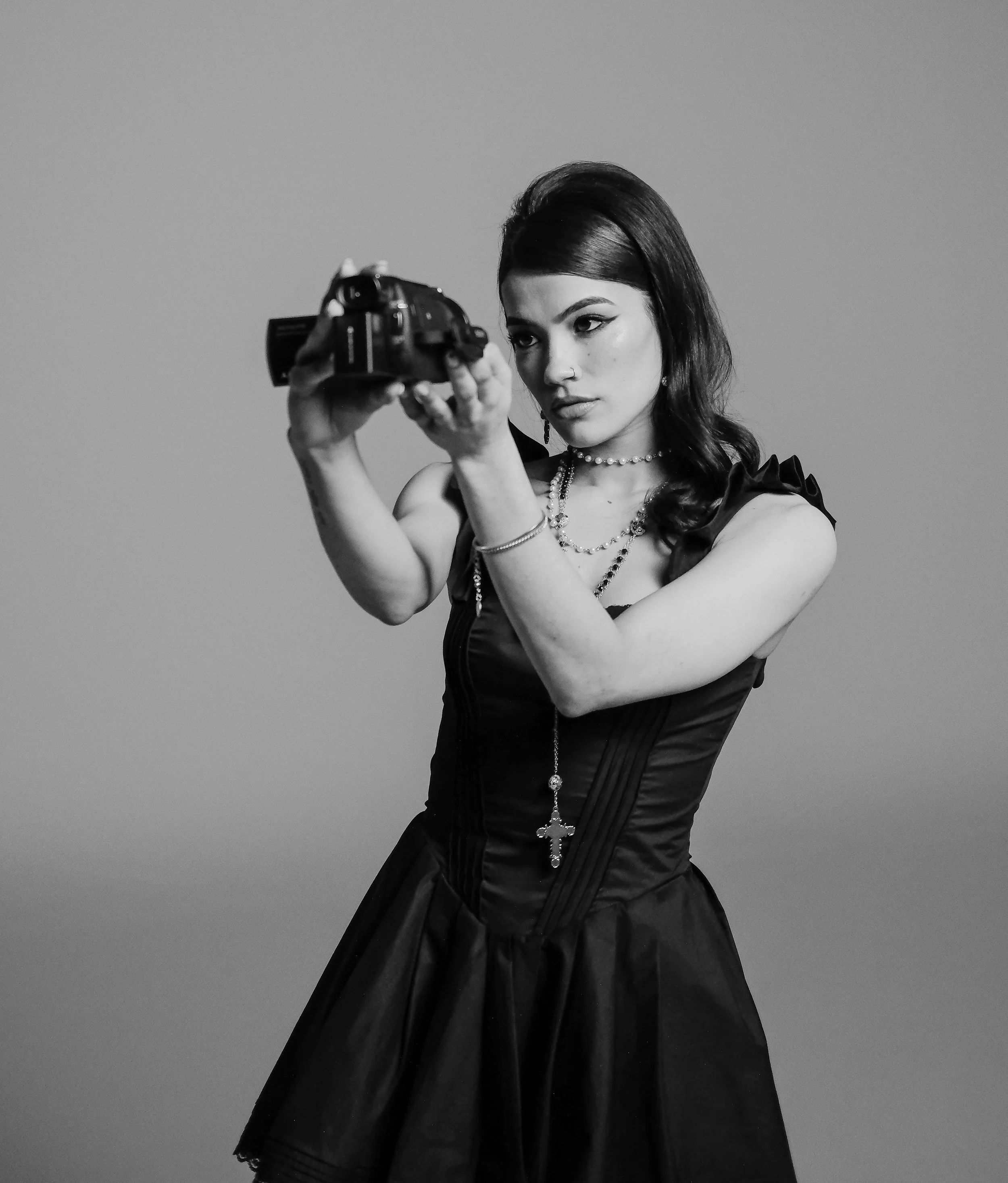 A woman holding a camera up to take a photo, dressed in a Gothic style with dark makeup, a black dress, and jewelry, in a studio with plain background.