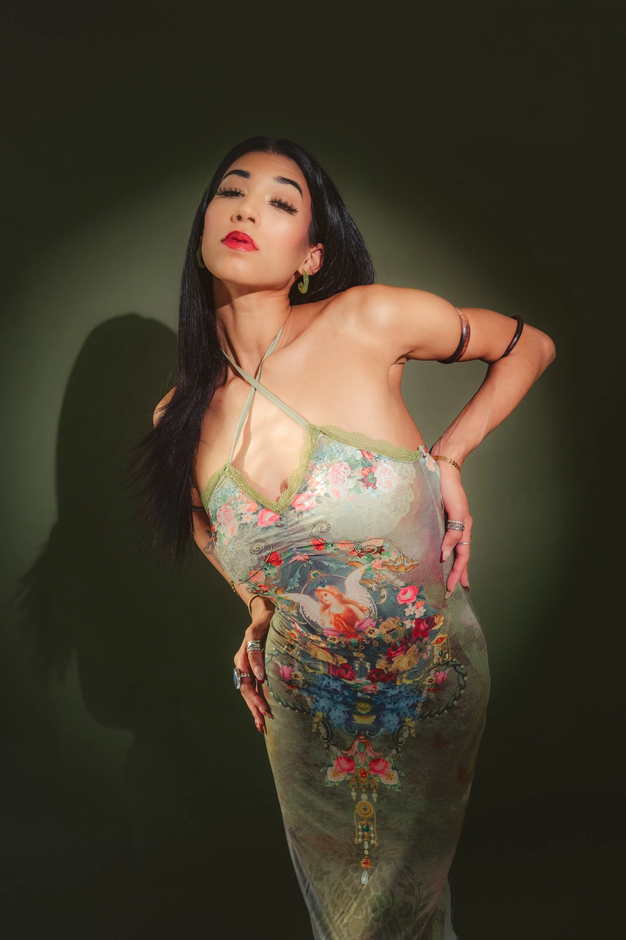 A woman with dark hair, red lipstick, and makeup posing in front of a dark green background wearing a colorful, patterned dress with a plunging neckline, showcasing floral and mythical design elements.